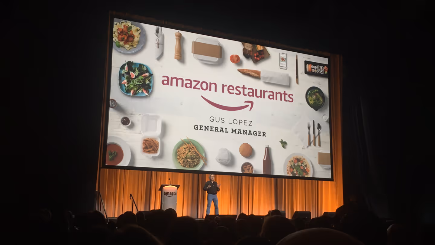 Presenter on stage at Amazon conference with large screen displaying Amazon Restaurants branding and food imagery.