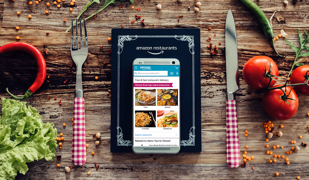 Smartphone displaying Amazon Restaurants app on menu board surrounded by fresh vegetables and utensils on wooden table.