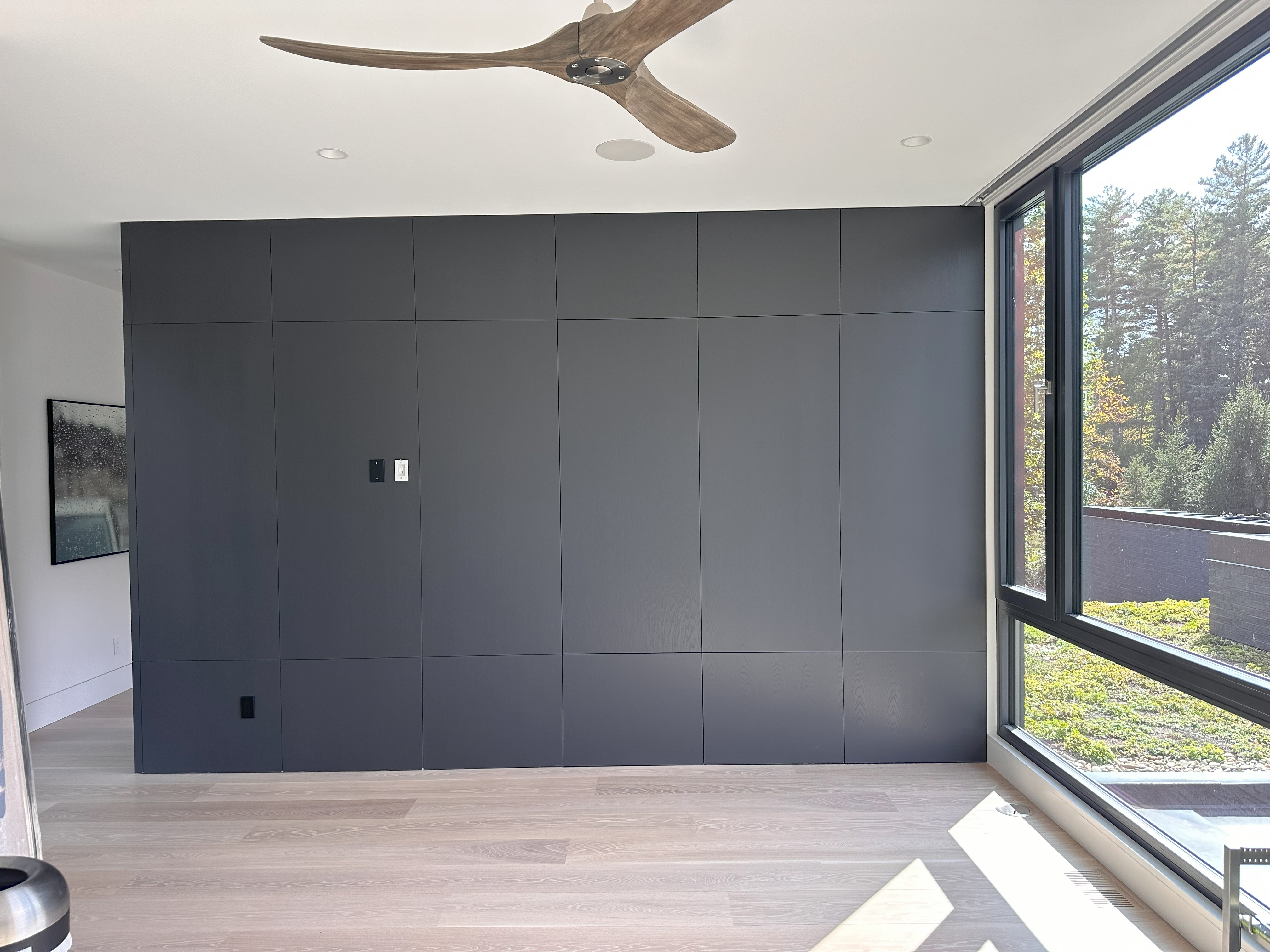 Modern room interior with a large dark gray paneled wall, light wooden floor, large floor-to-ceiling window showing outdoor greenery, a wooden ceiling fan with three blades, and a framed painting partially visible on the left wall.