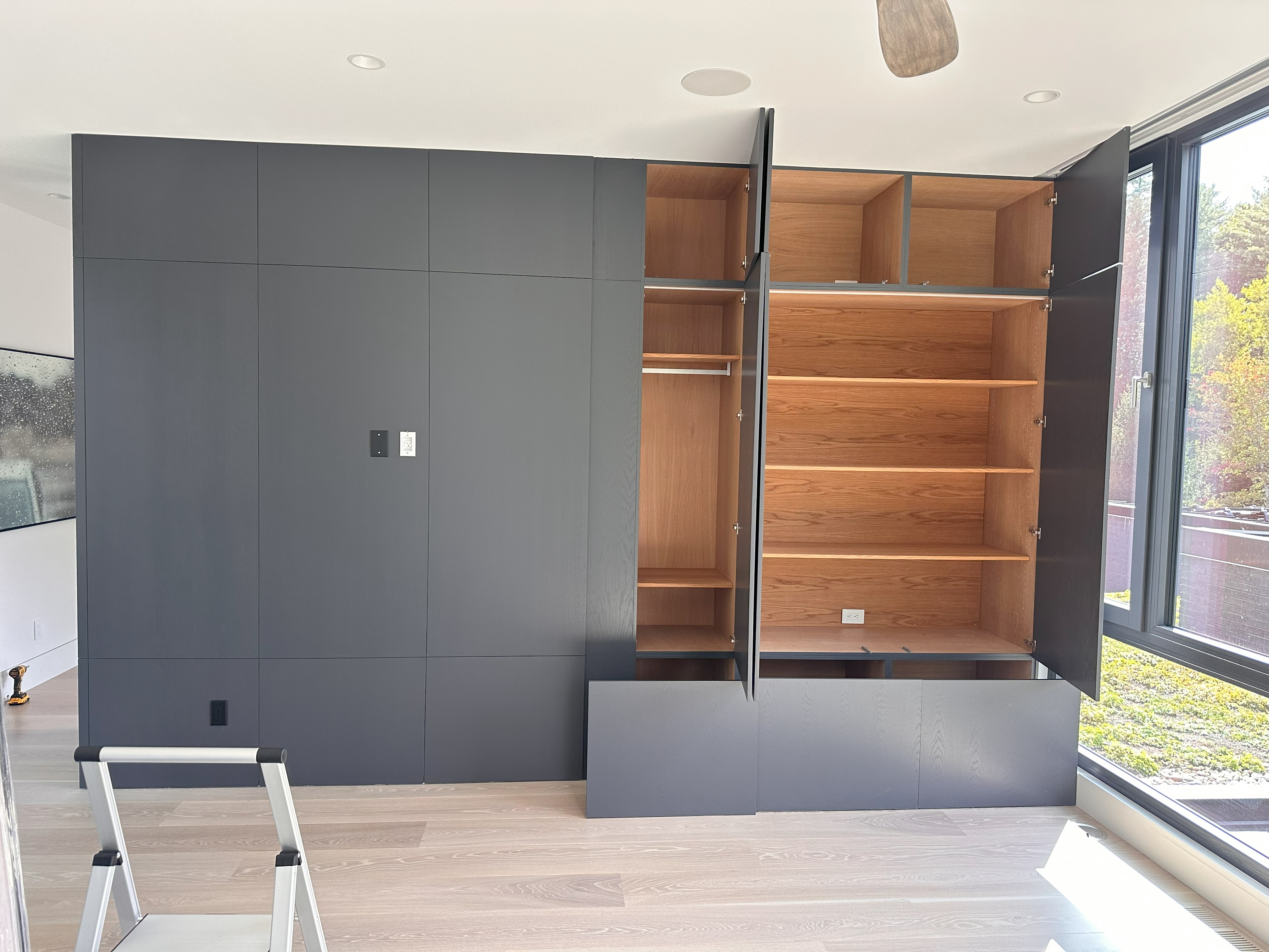 Modern built-in dark gray wall unit with open wooden shelves and compartments on the right, closed panels on the left; a small step ladder is in front, and large windows on the right show outdoor greenery.
