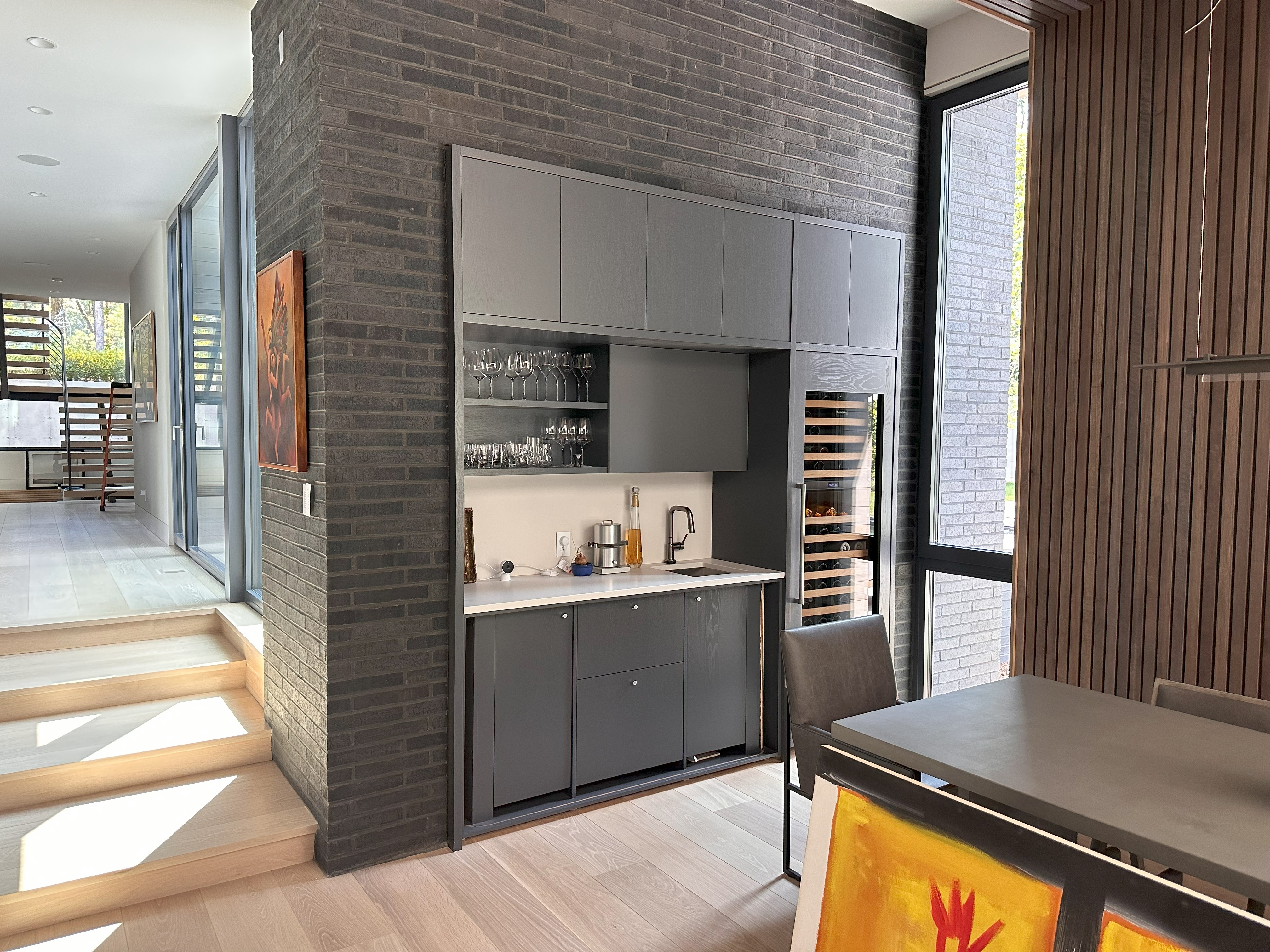 Modern kitchen area with dark gray cabinets, built-in wine coolers, sink with black faucet, glassware on shelves, and wooden dining table with gray chairs next to large windows letting in natural light.