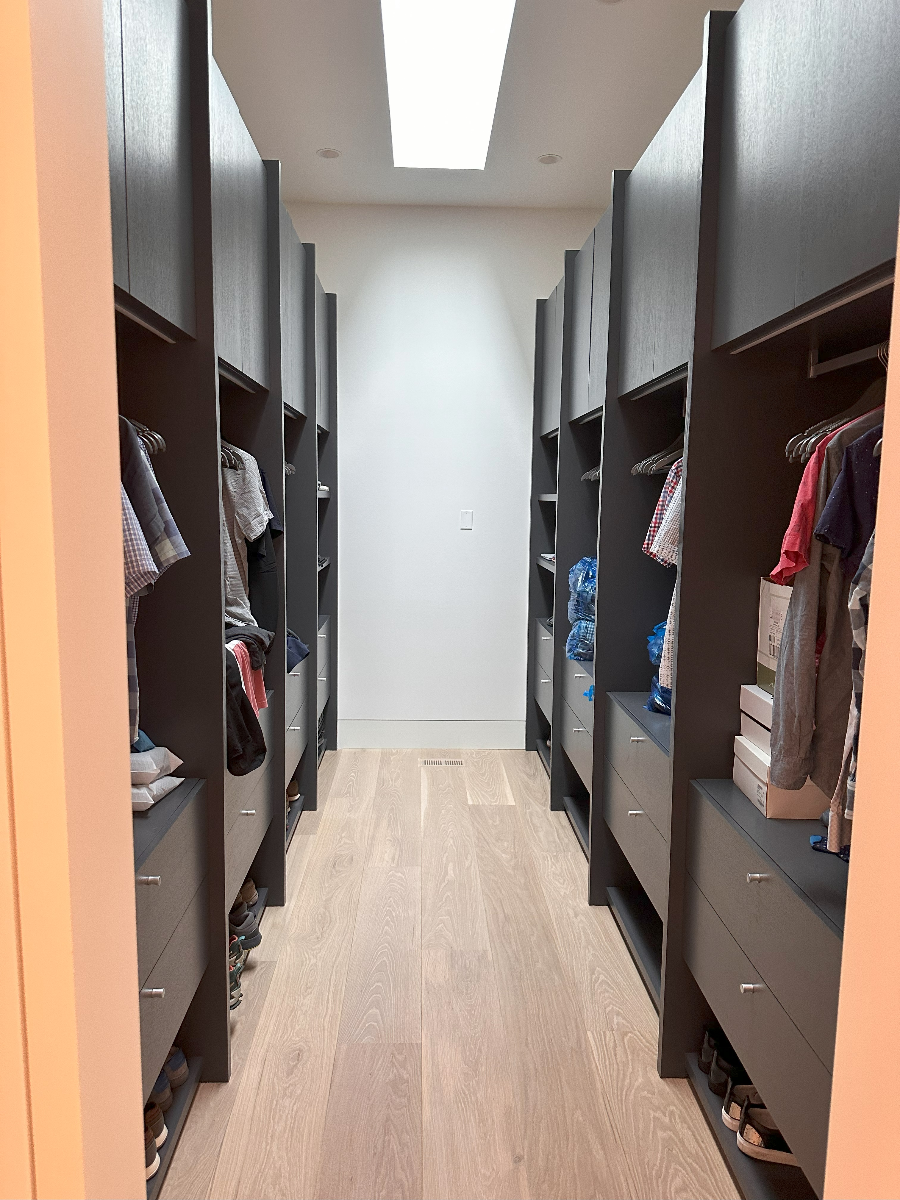 Walk-in closet with dark wooden shelves and drawers on both sides, clothes hanging on rods, shoes on lower shelves, light wood flooring, and a rectangular skylight on the ceiling.