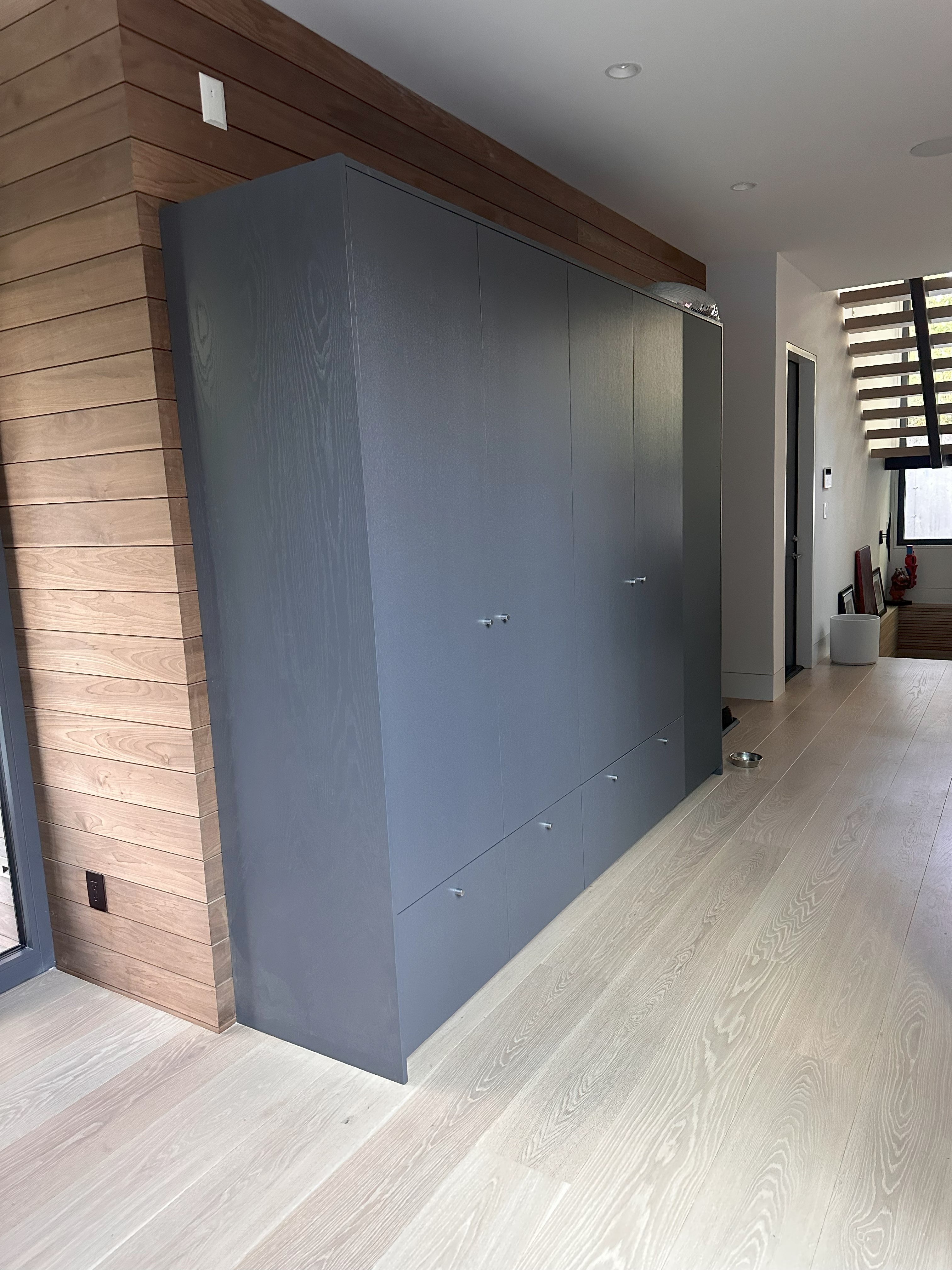 Modern hallway with light wood flooring and a large gray wardrobe against a wood-paneled wall, next to stairs and a small door.