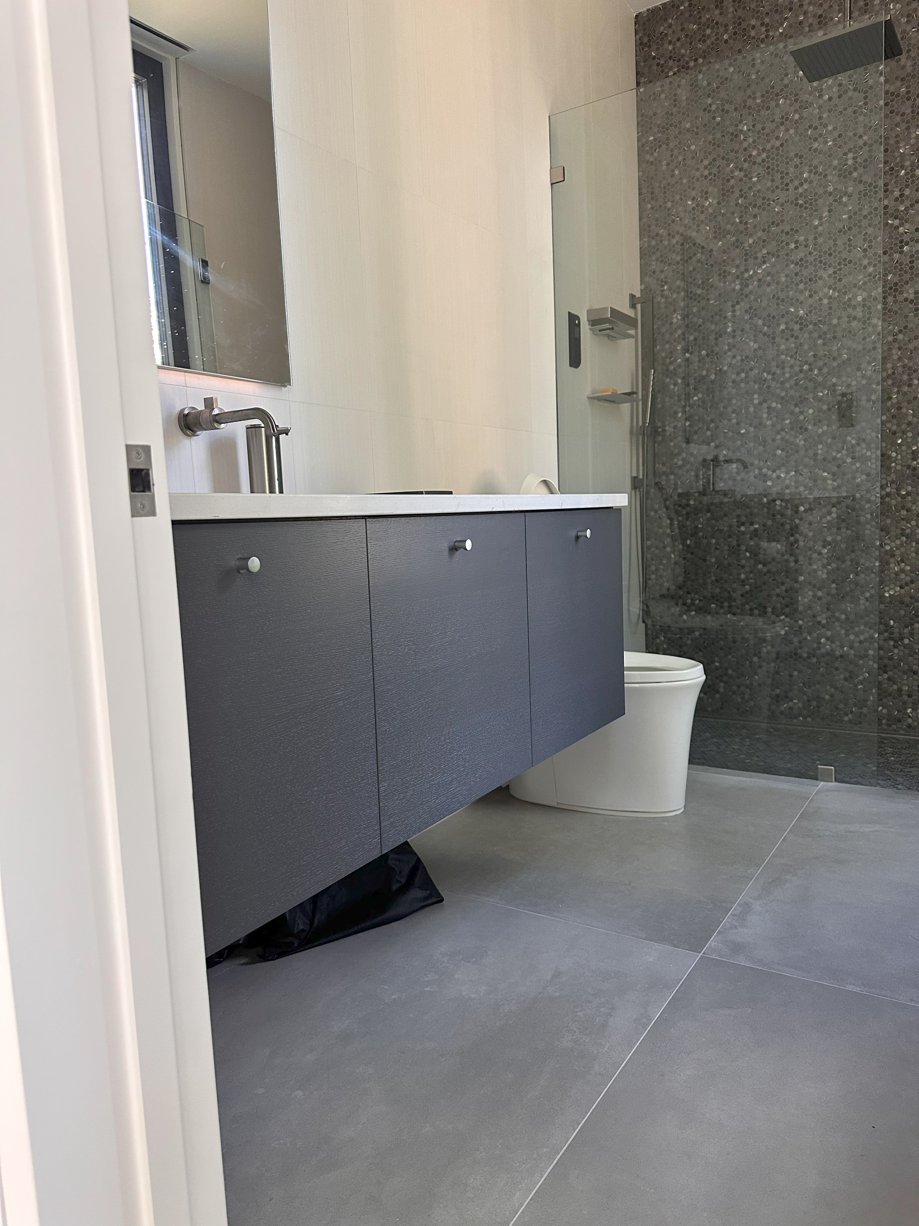 Modern bathroom interior with gray floating vanity, white countertop, wall-mounted faucet, mirror, white toilet, and glass-enclosed shower with pebble tile wall and overhead showerhead.