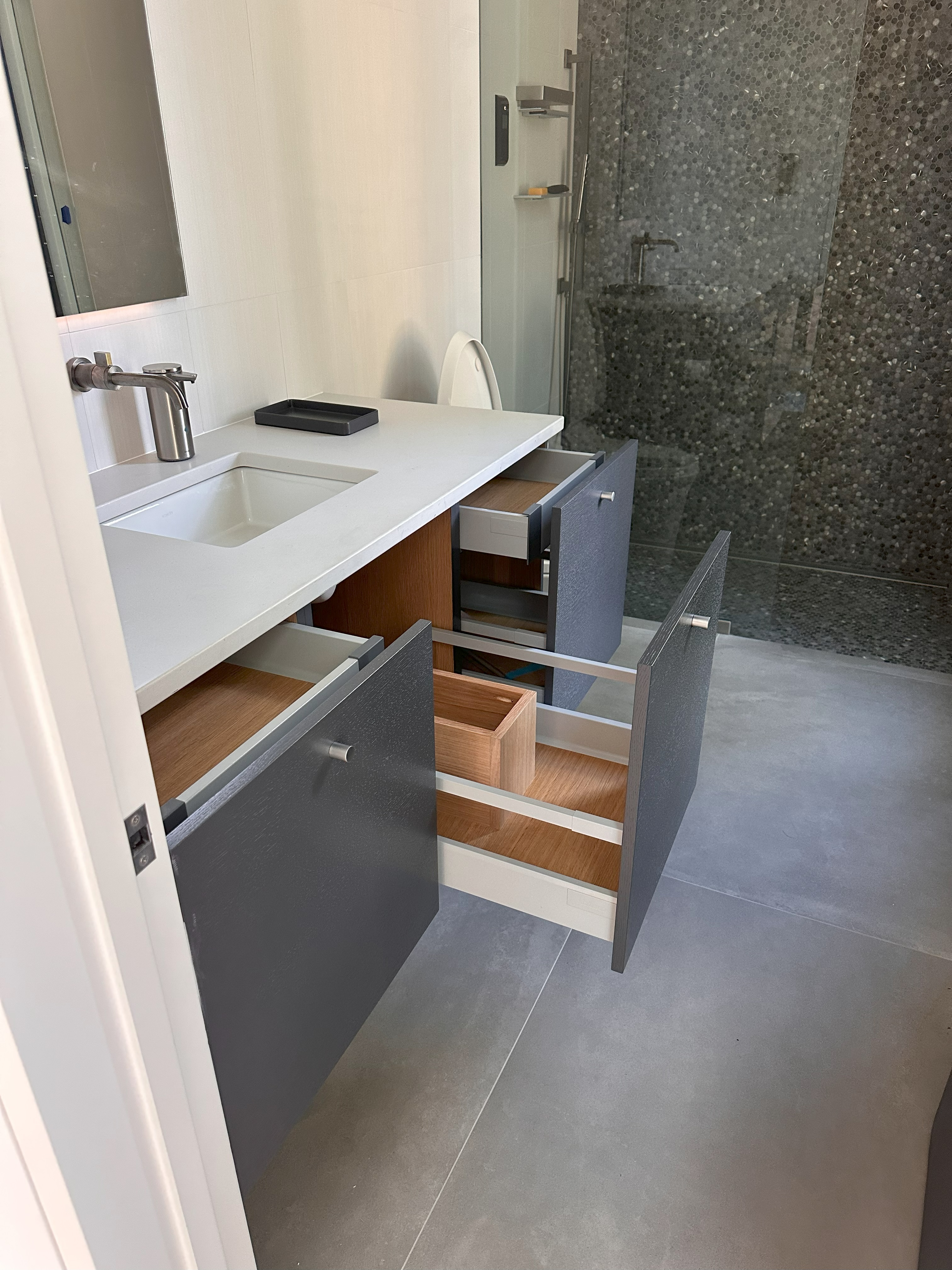 Modern bathroom vanity with white countertop, built-in rectangular sink, stainless steel faucet, and open gray cabinet drawers displaying wooden interiors, next to a shower area with a pebble-patterned glass wall.