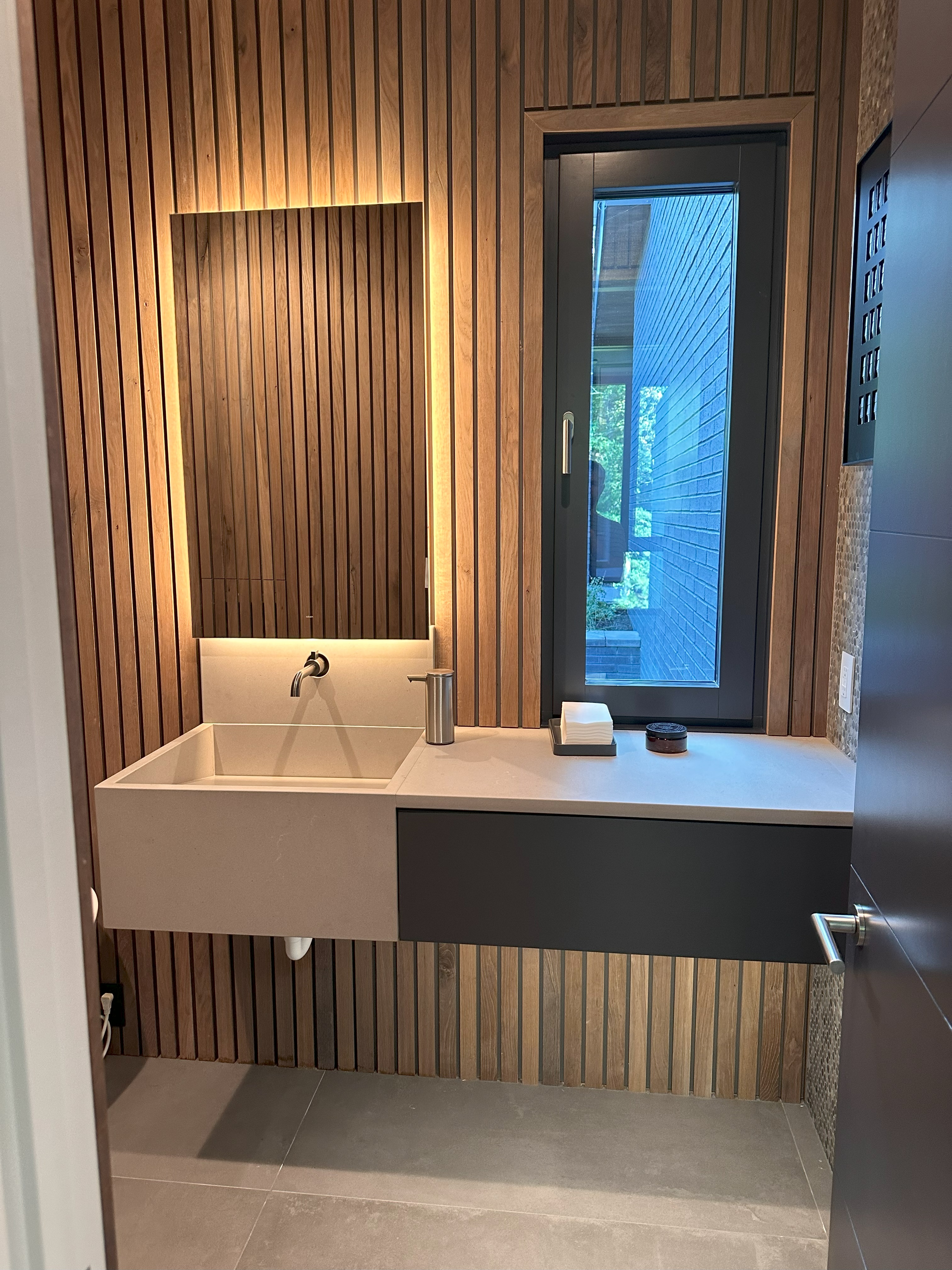 Modern bathroom with vertical wooden slat wall, rectangular sink with wall-mounted faucet, illuminated mirror, dark gray cabinet drawer, window, and door with metal handle.