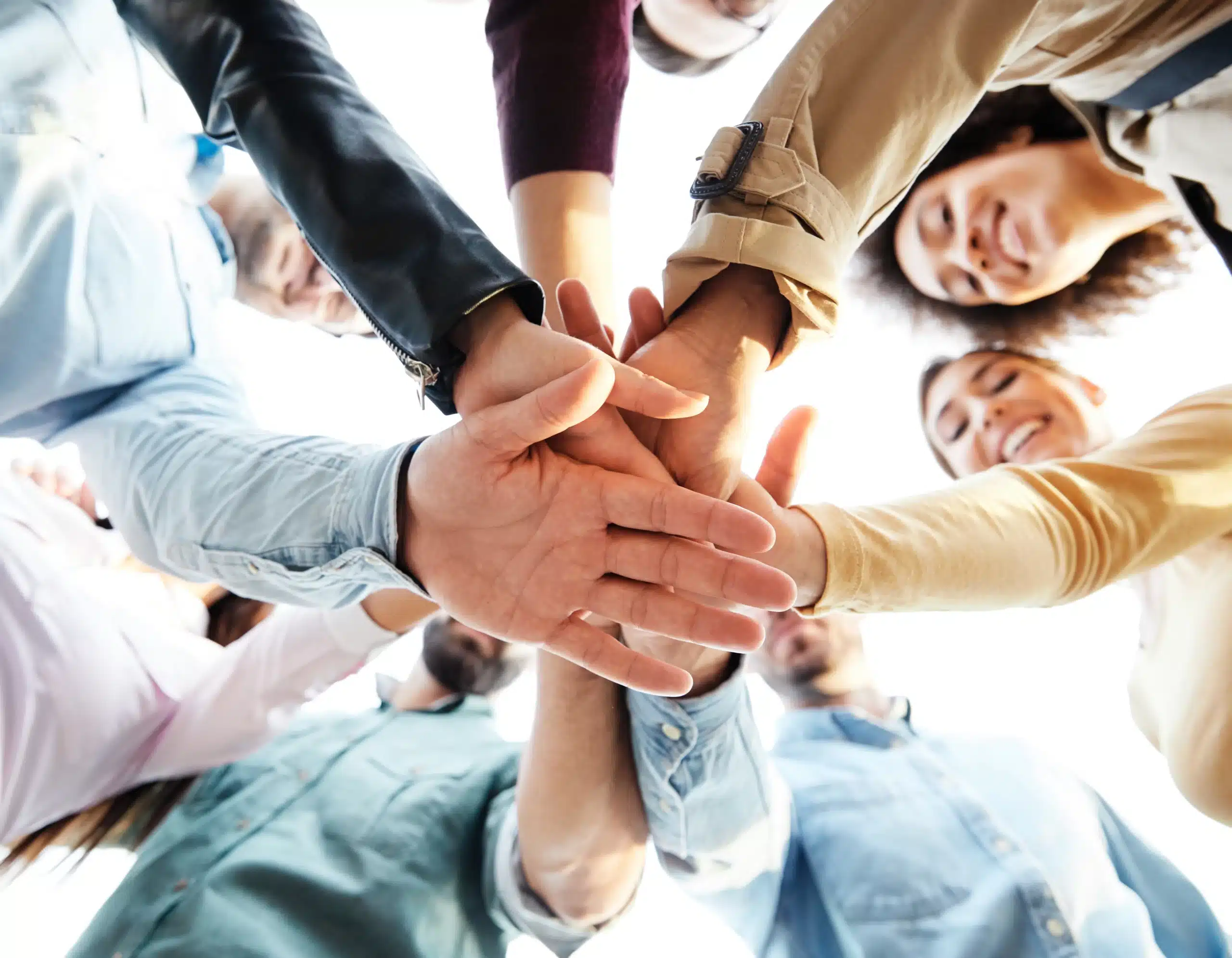 Group of people stacking hands together in a teamwork gesture, smiling from below.
