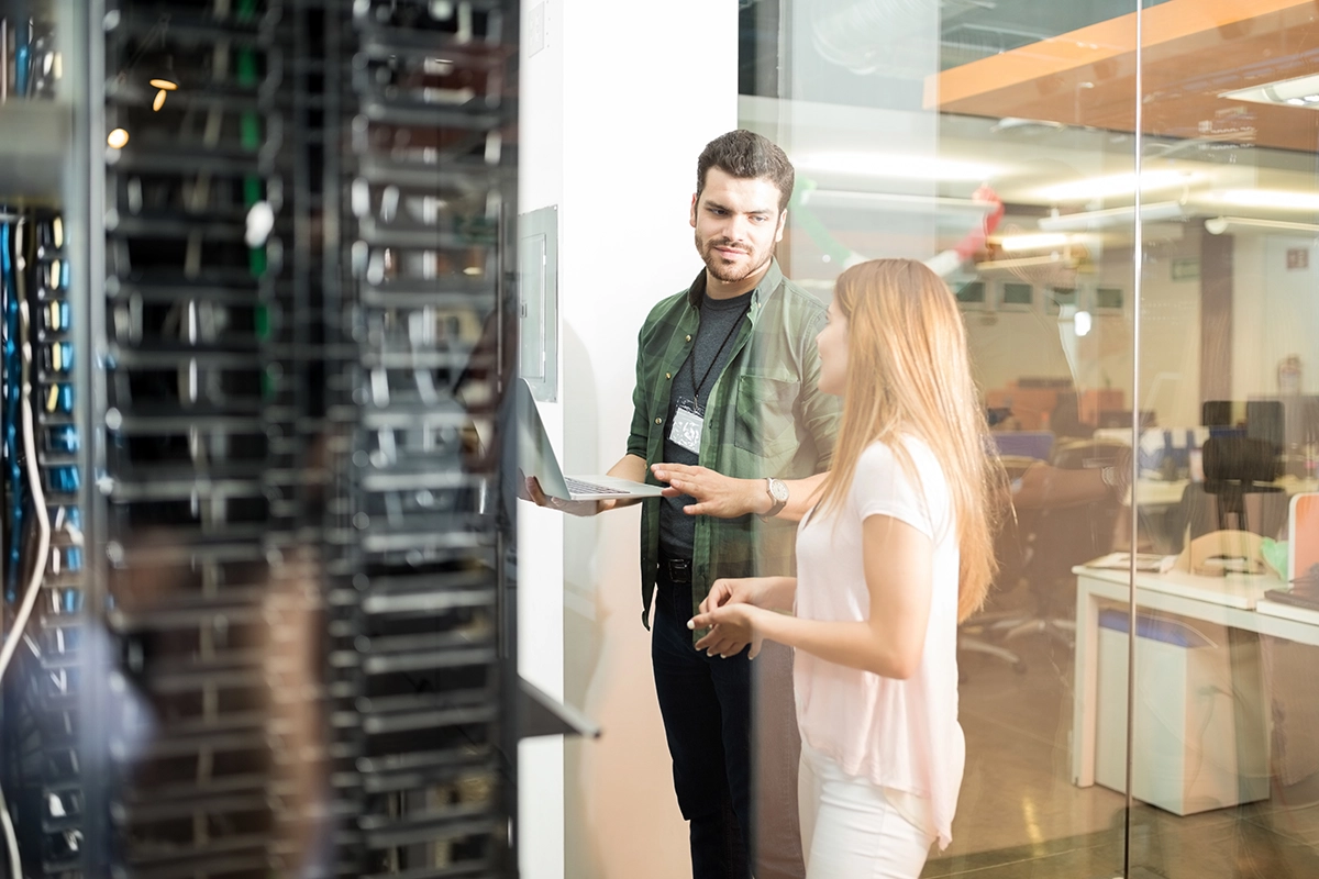 Two people in a server room, discussing something while looking at a laptop, with server racks visible.