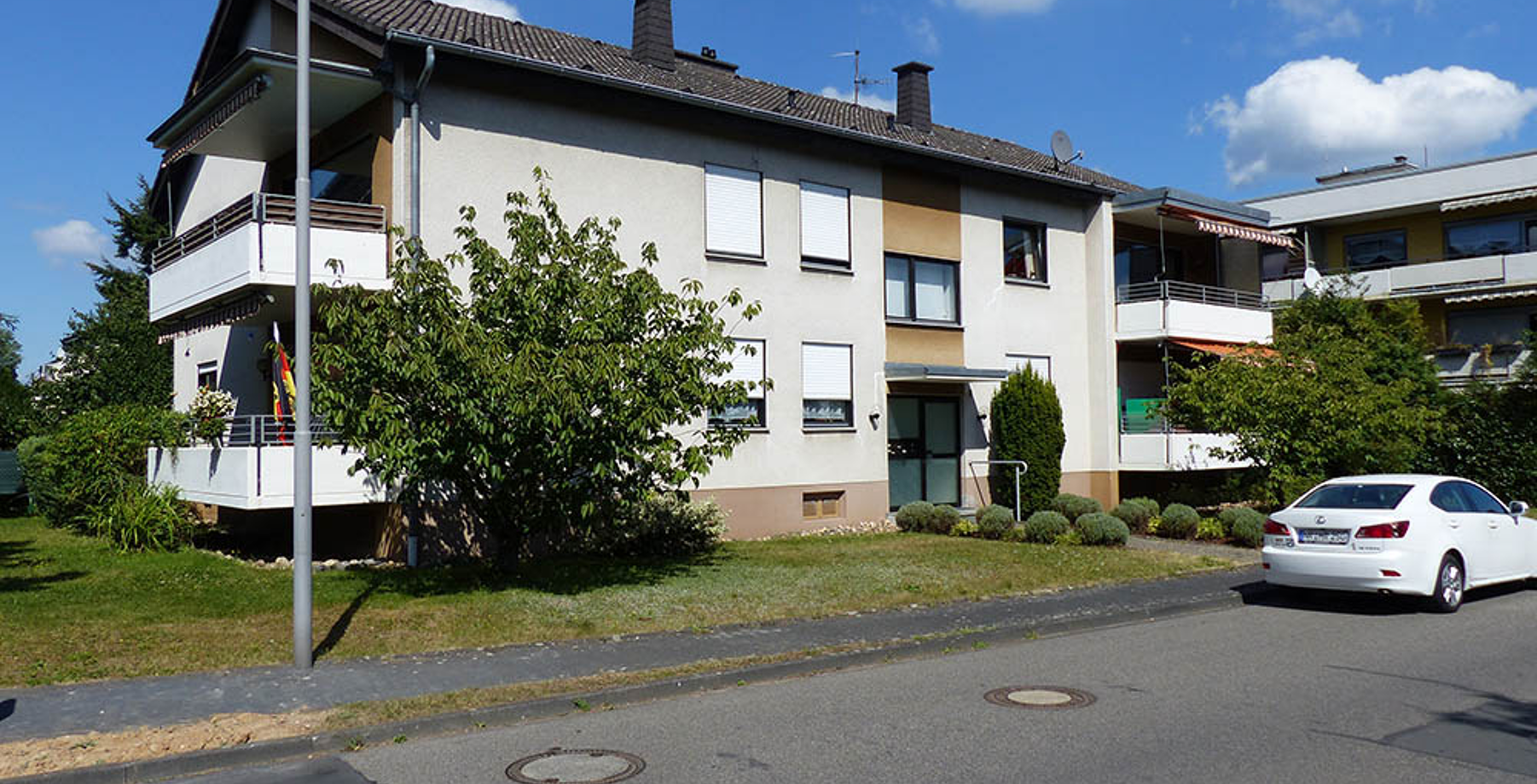 Two-story residential building with white balconies, green bushes, and a white car parked on the street.