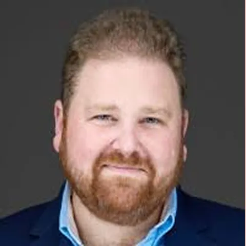 Smiling man with red hair and beard wearing a blue collared shirt and dark blazer against a gray background.