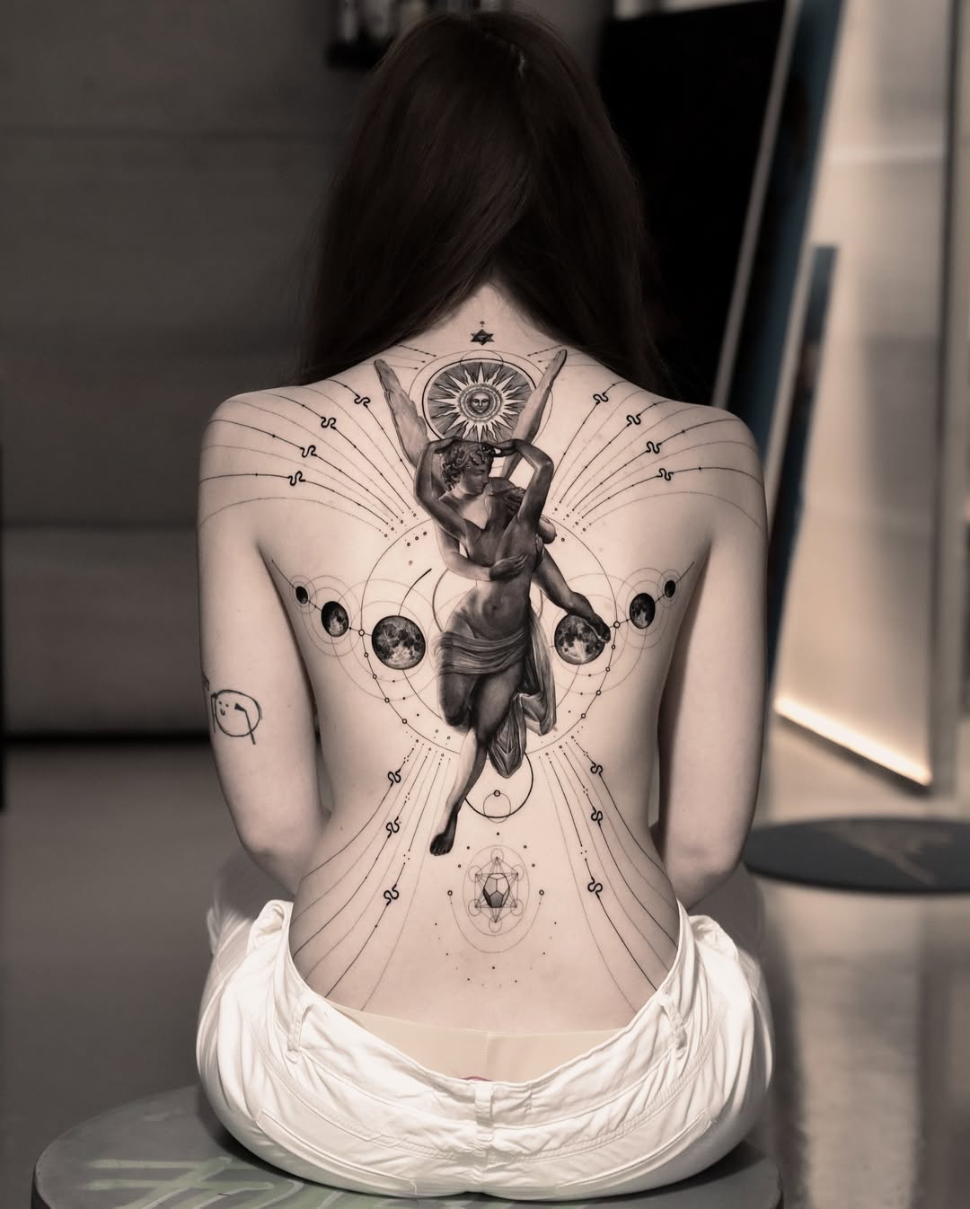 Frau mit Tattoos an Arm und Oberkörper posiert mit hochgezogenem linken Arm, trägt dunkle Hose und hält sich die Brust.