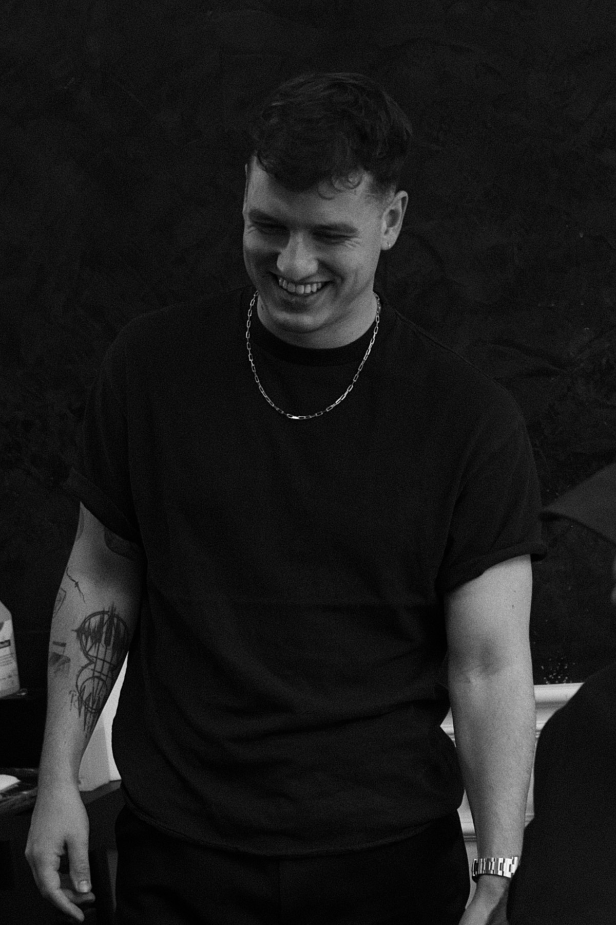 Junger Mann mit kurzem Haar, lächelnd, trägt eine Kette, ein Armband und ein schwarzes kurzärmeliges Hemd mit tätowiertem rechten Arm.