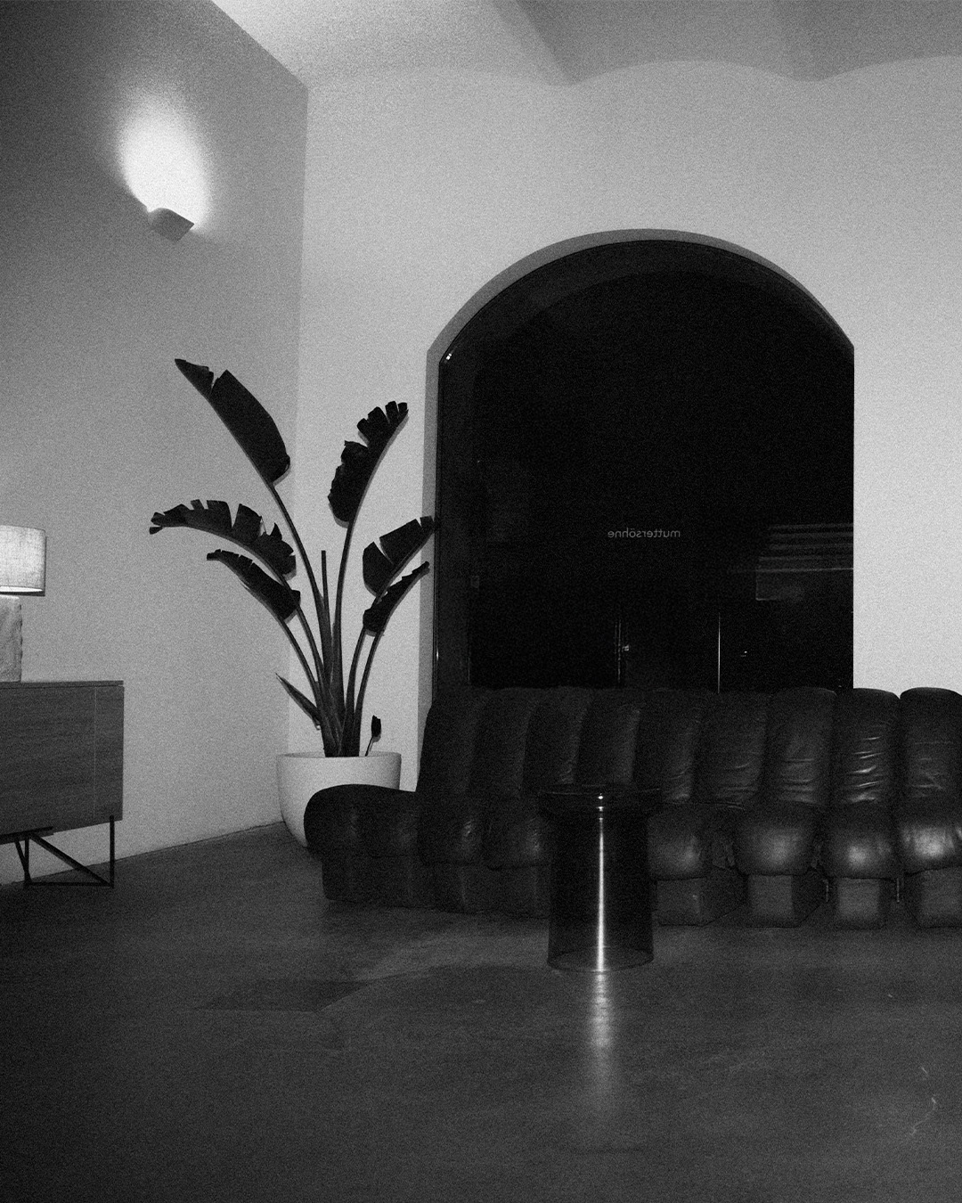 Schwarz-weiß-Foto eines modernen Wohnzimmers mit einer großen Ledercouch, einem runden Glastisch, einer Zimmerpflanze in einem weißen Topf und einem beleuchteten Lampenschirm auf einem Sideboard.