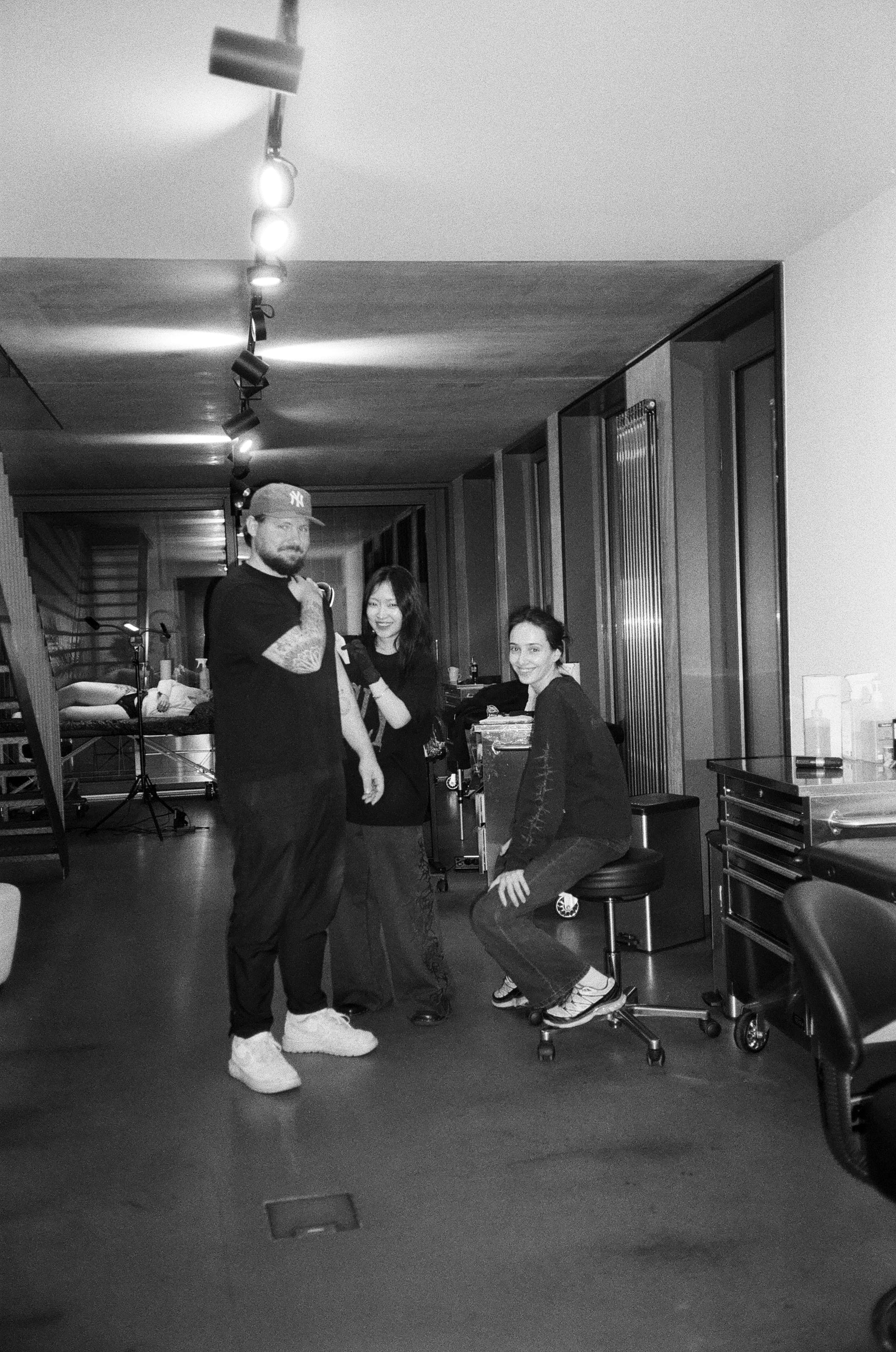 Schwarzweißfoto eines Mannes mit Baseballkappe, der sich tätowieren lässt, begleitet von zwei lächelnden Frauen in einem modernen Studio.