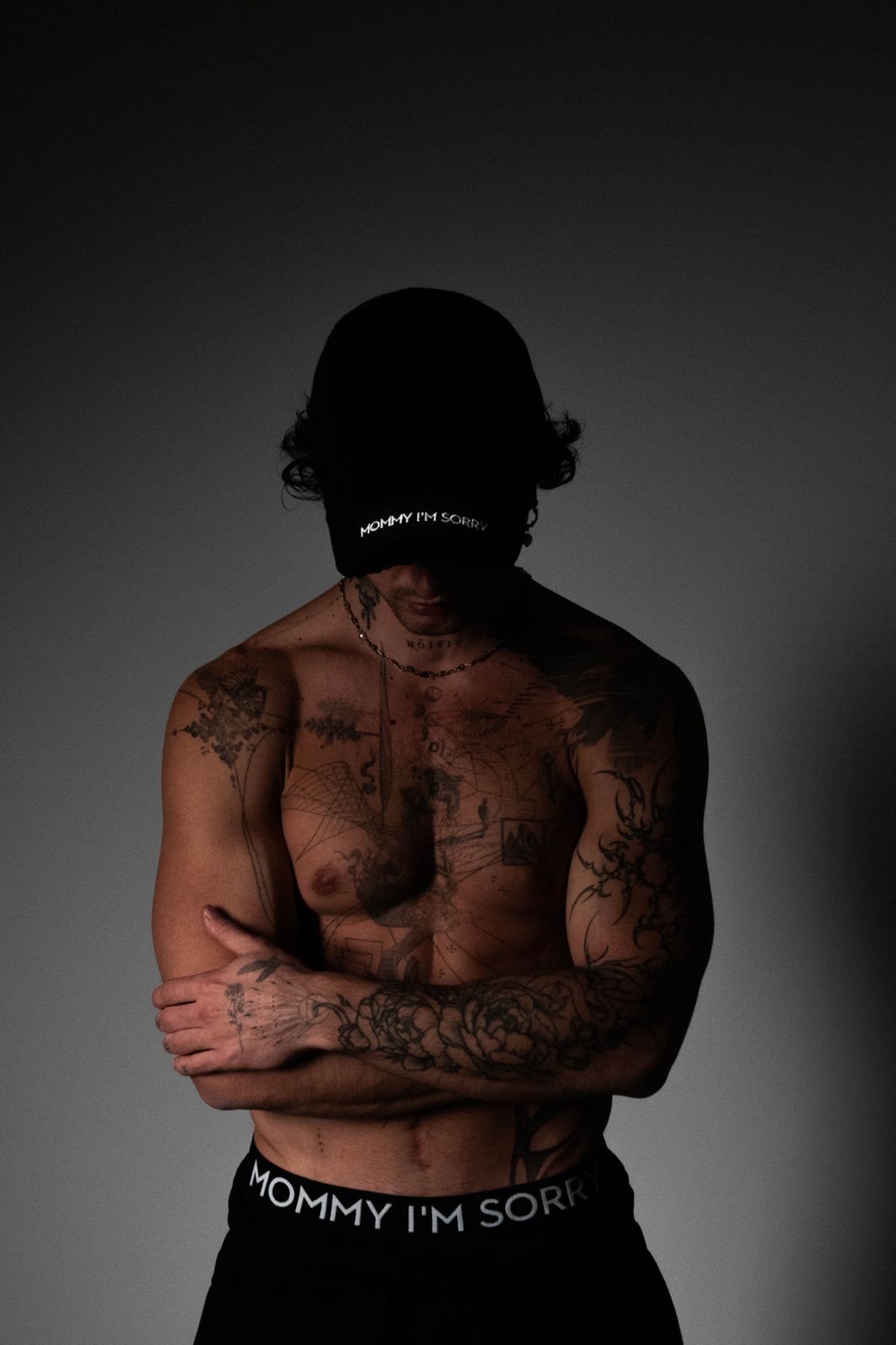 Muskulöser, tätowierter Mann mit schwarzer Kappe und Hose, auf denen 'Mommy I'm Sorry' steht, mit verschränkten Armen vor dunklem Hintergrund.