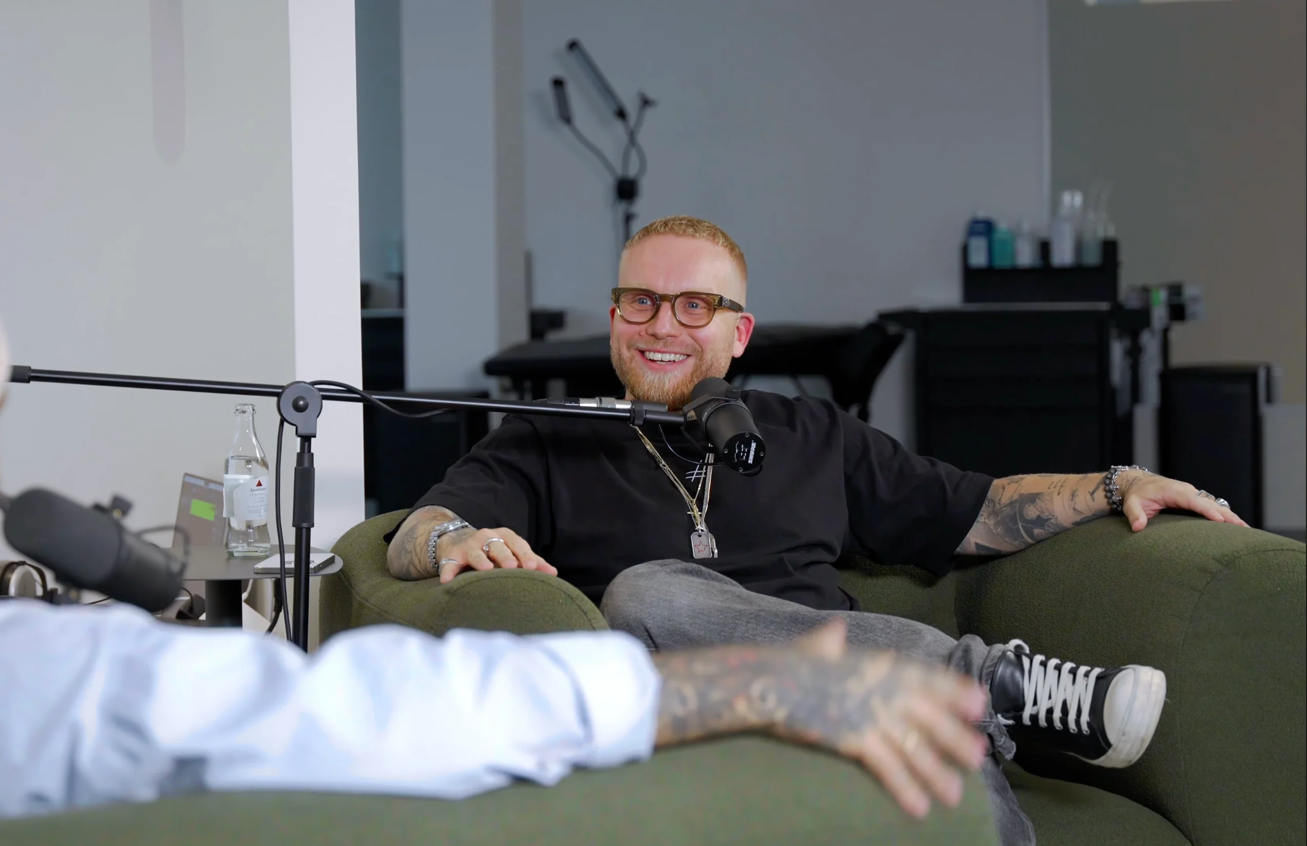 Lächelnder Mann mit Brille und Tätowierungen sitzt auf einem gemütlichen grünen Sofa und spricht in ein Mikrofon.