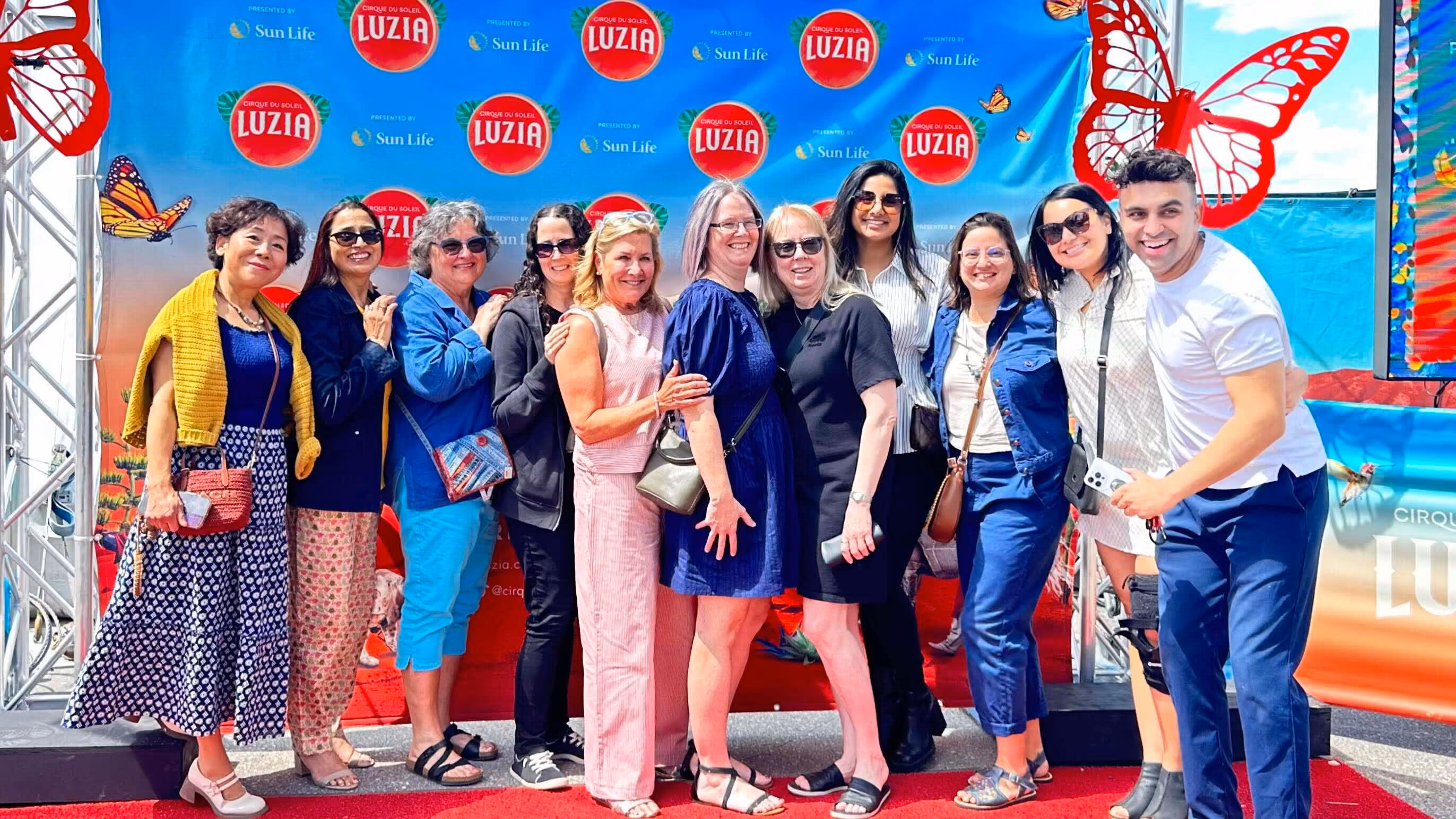 Group of ten people posing on a red carpet with Cirque du Soleil Luzia backdrop and butterfly decorations.