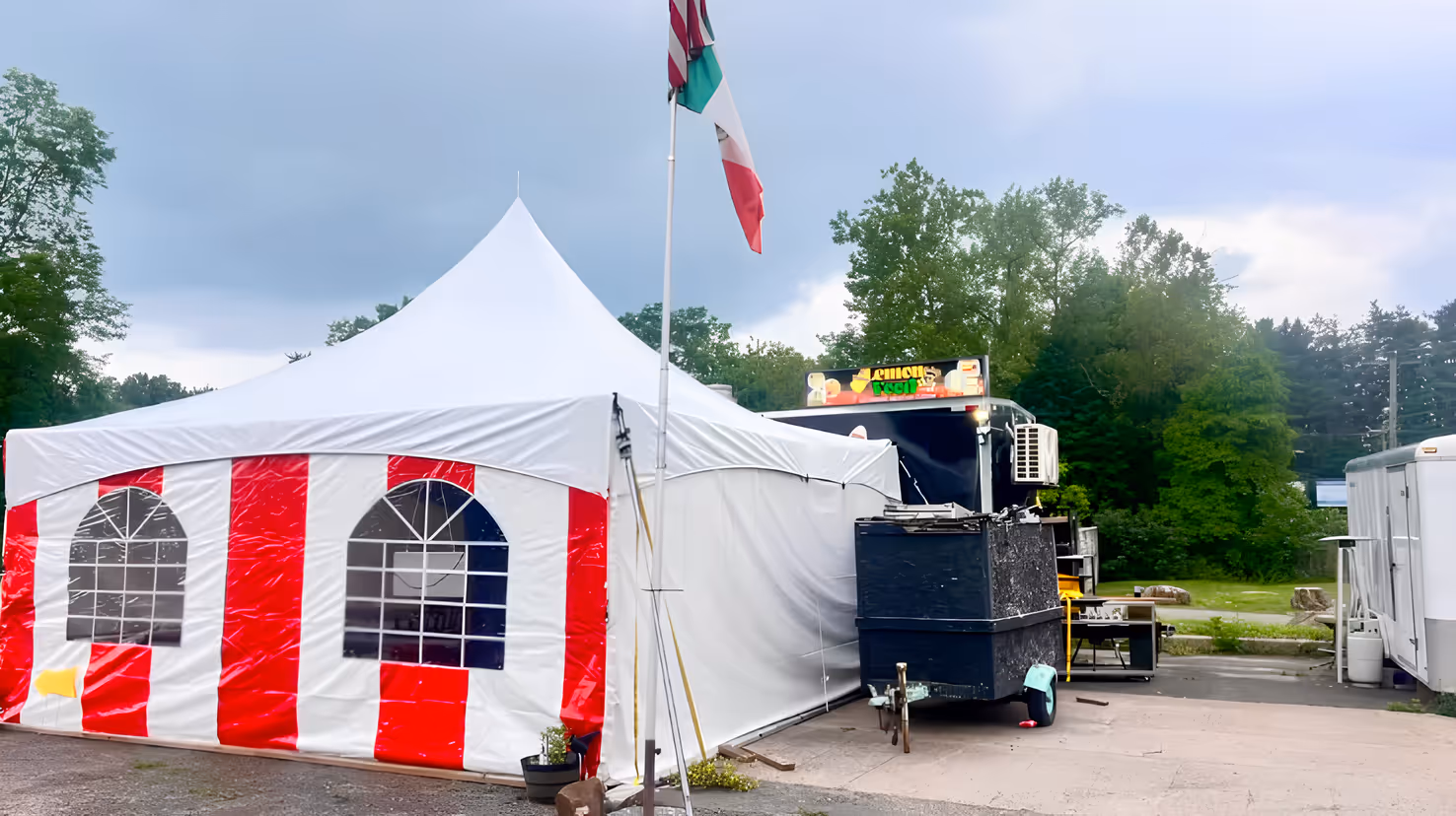 A white and red striped tent stands under a cloudy sky, with a Mexican flag waving above. Nearby, a food truck.