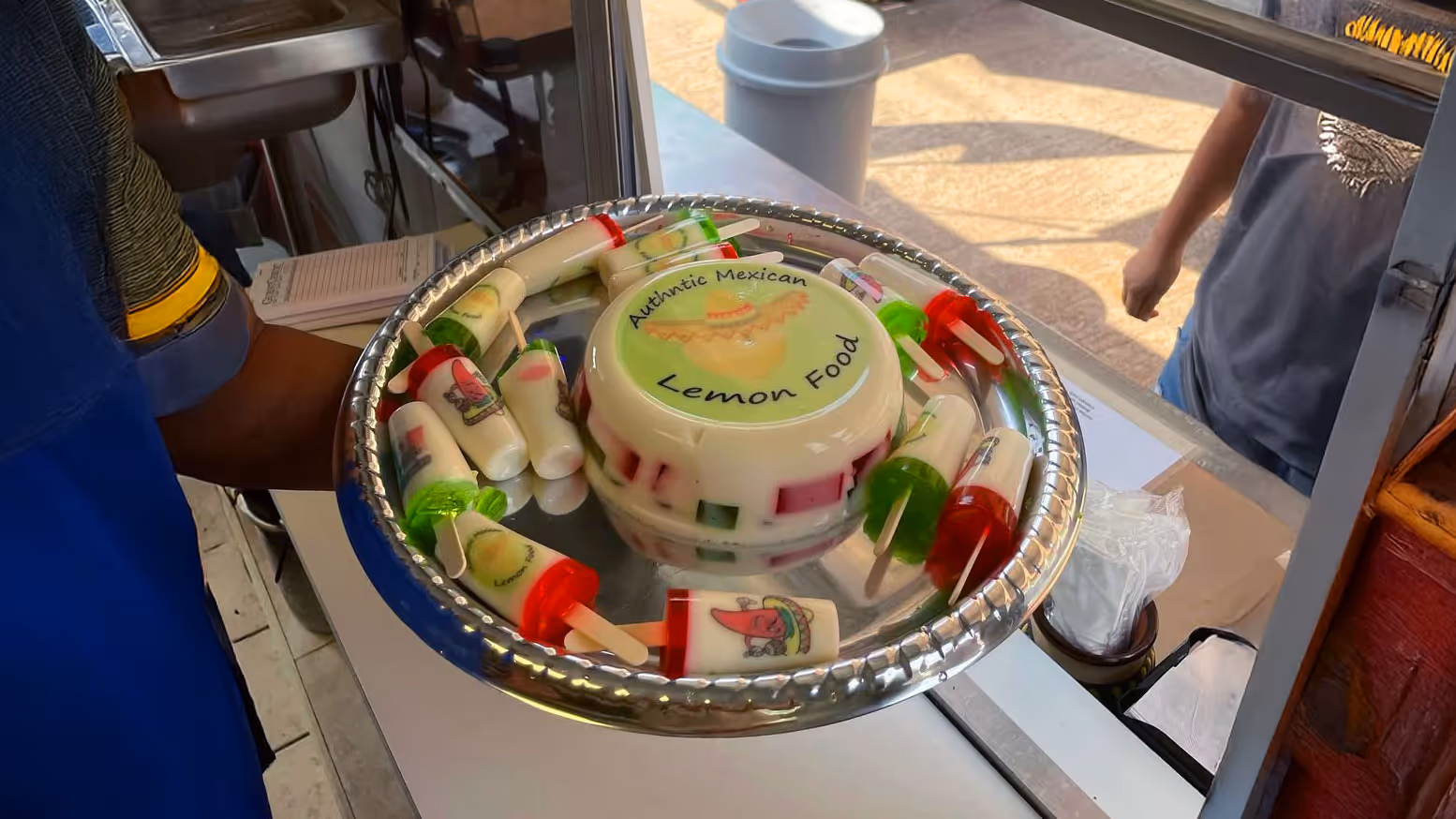 A tray with assorted vibrant Mexican treats, including colorful jello popsicles and a jiggly Mexican jello labeled "Authentic Mexican Lemon Food". The jello at the center is held by a vendor at a food truck.