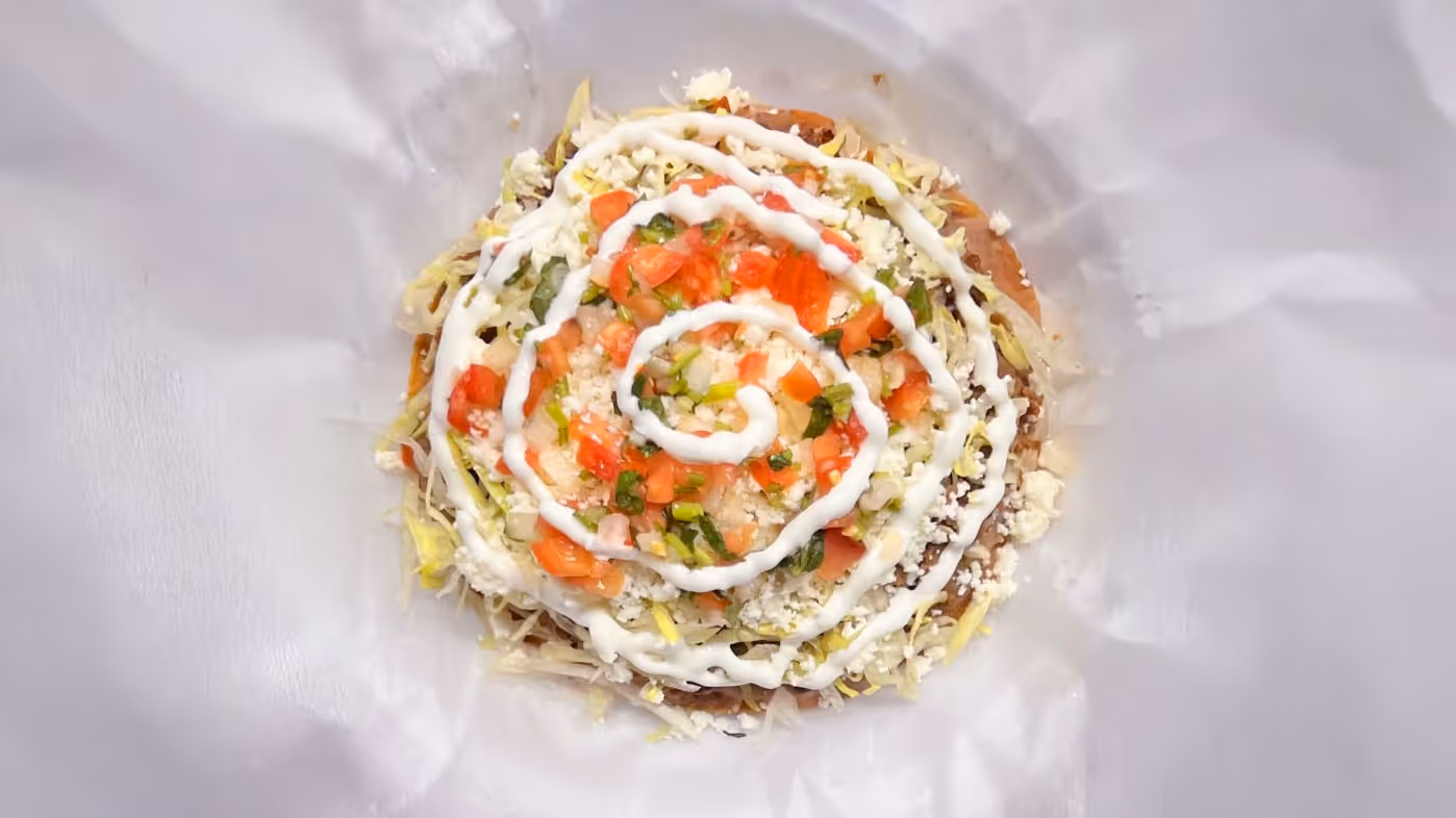 A vibrant Mexican tostada is topped with shredded lettuce, diced tomatoes, cheese, and a spiral drizzle of sour cream. It sits on white parchment paper.