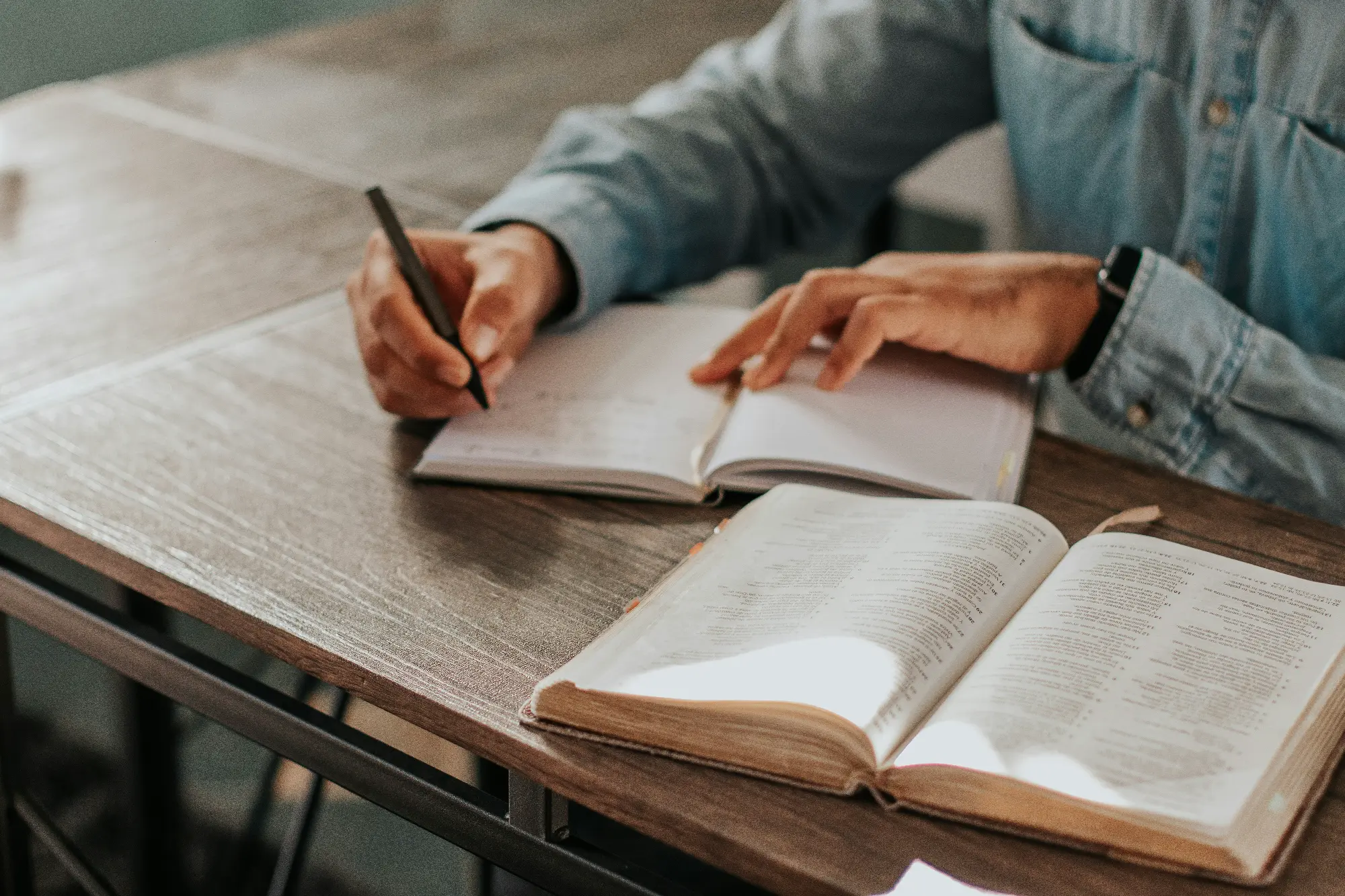 A person writing in a notebook while referencing an open Bible on a wooden table.