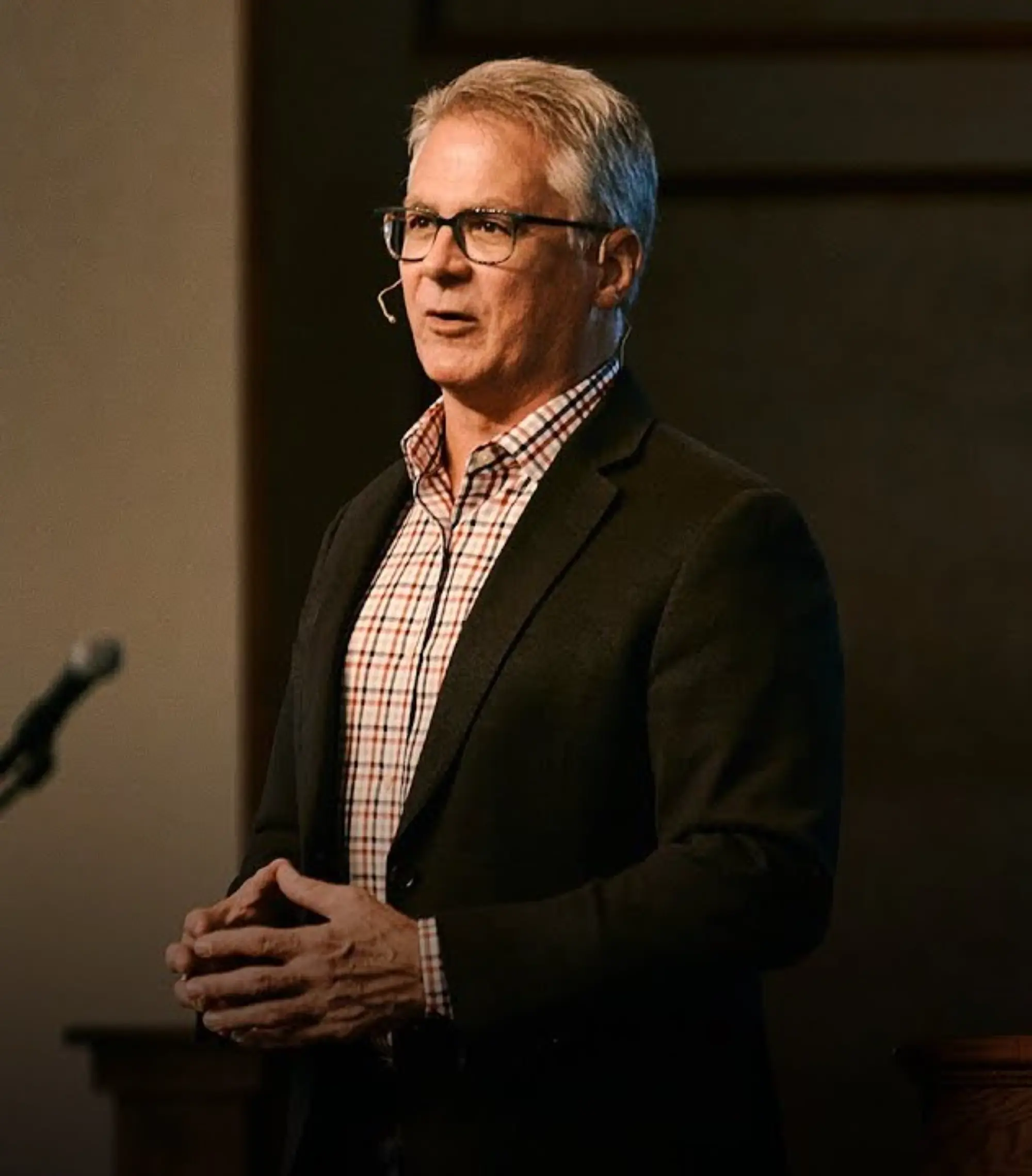 A pastor in a plaid shirt under a blazer speaks to an audience, with a microphone visible nearby, in a warm, indoor setting.
