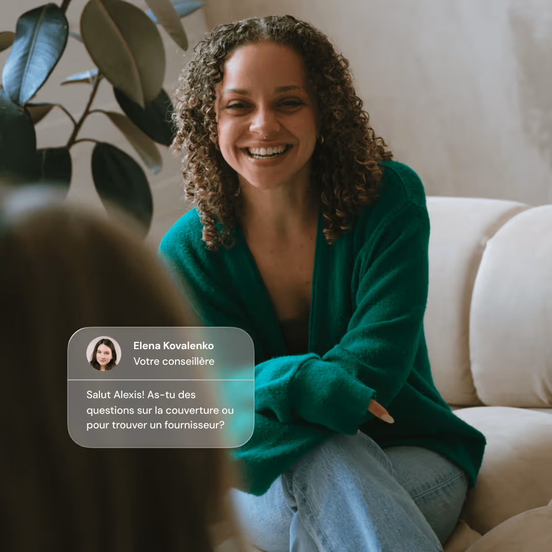 Smiling woman with curly hair in a green sweater sitting on a couch, talking to another person.