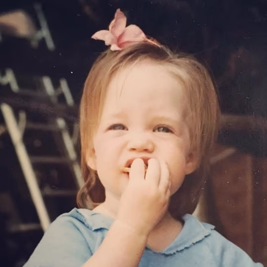 Young child with light hair and a pink flower clip eating food while wearing a light blue shirt.