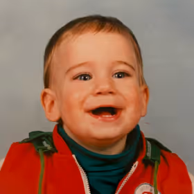 Smiling toddler with short hair wearing a red jacket and teal turtleneck against a gray background.