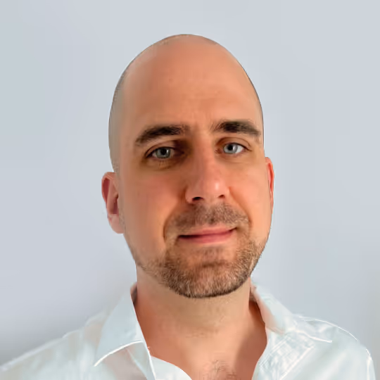Portrait of a bald man with light beard wearing a white shirt against a light gray background.