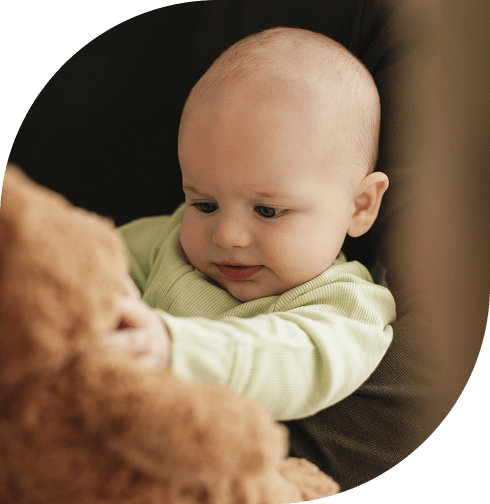 Baby in green outfit looking at and holding a brown teddy bear while seated in a dark chair.