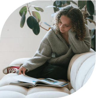 Curly-haired woman in a gray sweater reading a large book while sitting on a white couch.