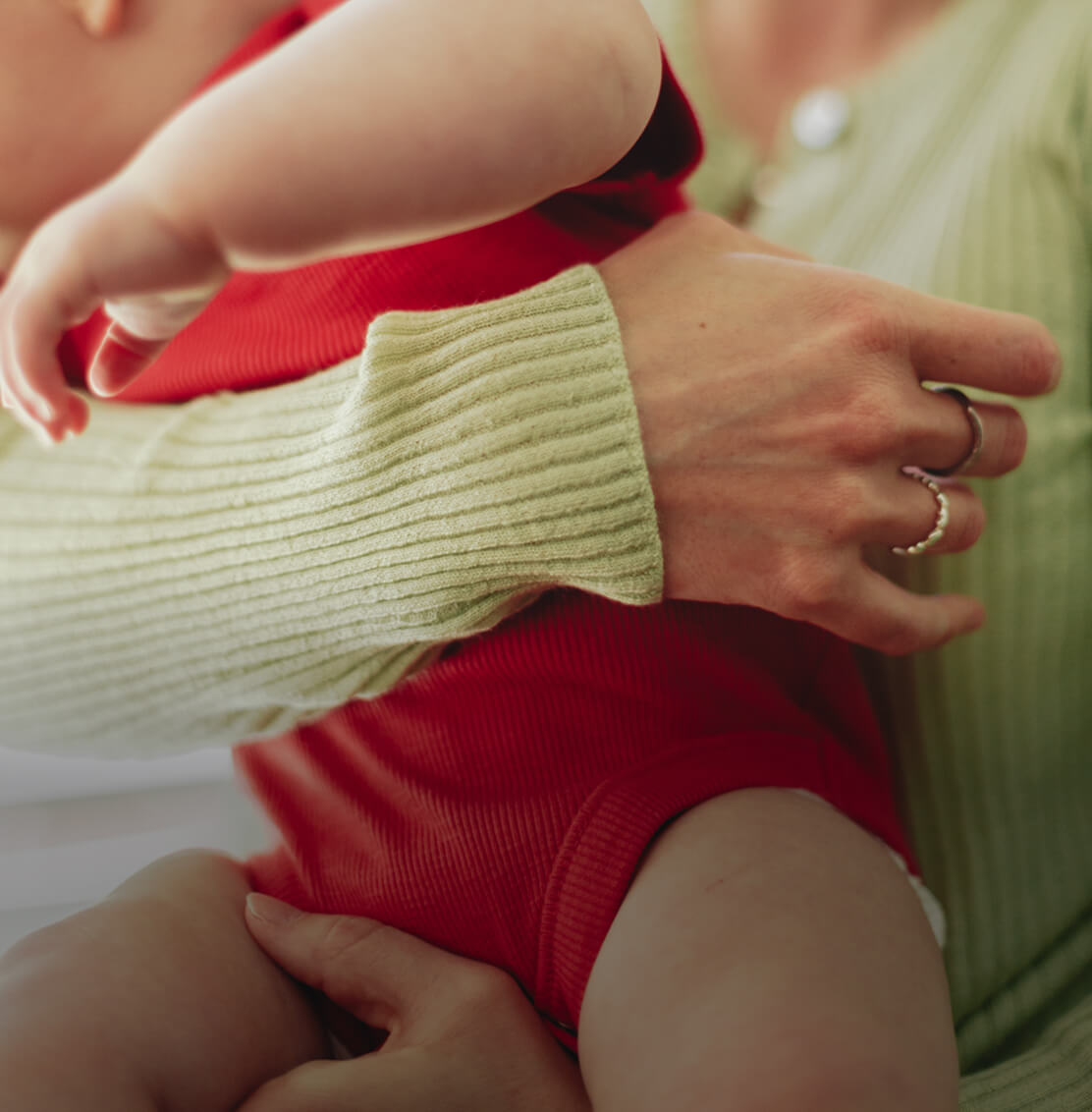 Close-up of an adult holding a baby in a red outfit, adult wearing green ribbed sweater and rings.