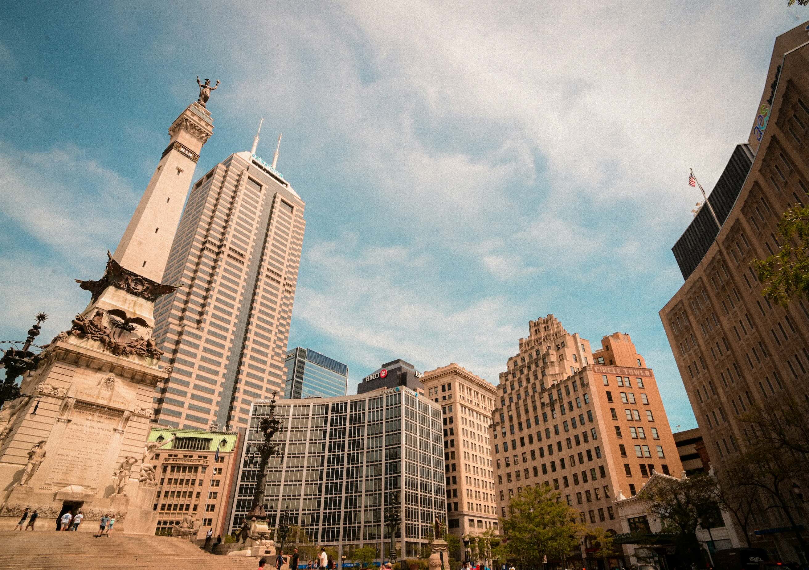 Monument Circle in downtown Indianapolis with the Soldiers and Sailors Monument in the foreground, surrounded by Circle Tower and modern office buildings under a partly cloudy blue sky.