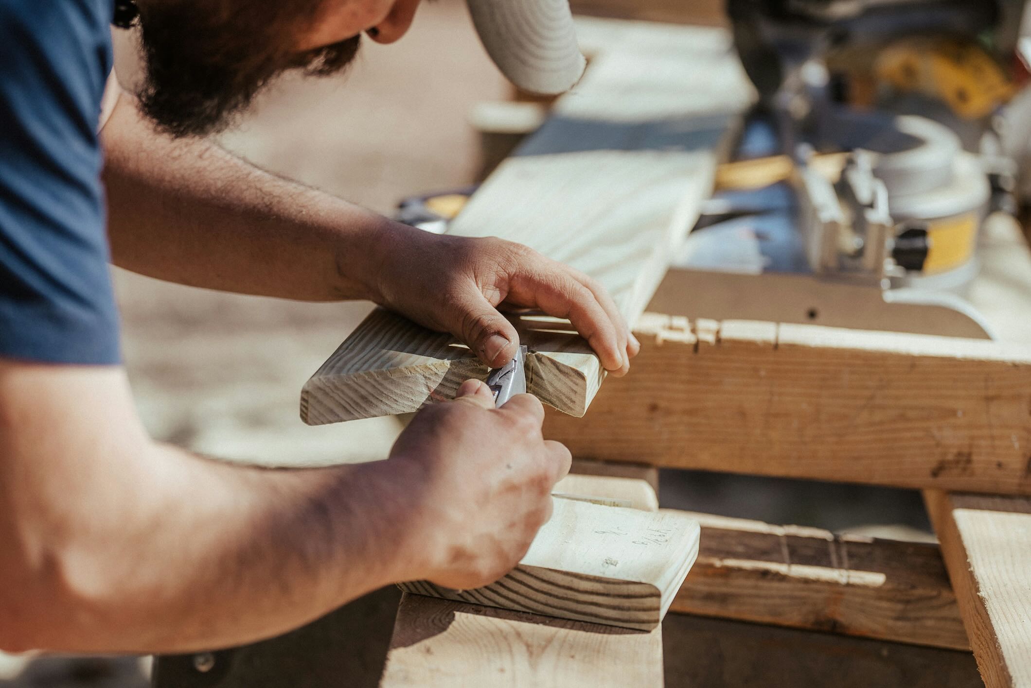 Close-up of a carpenter scoring a notch in a board with a utility knife on a jobsite, power tools blurred in the background.