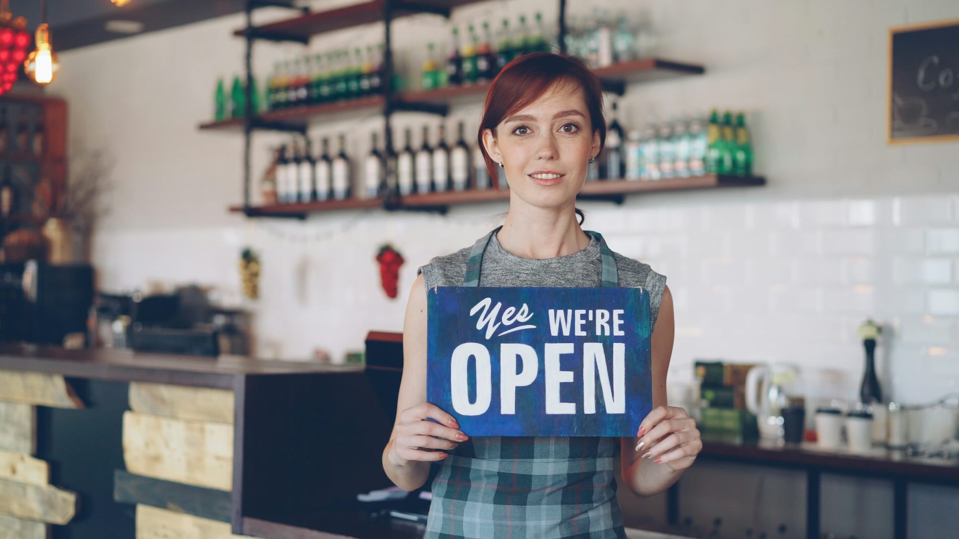 Small business owner in an apron holding a blue “Yes, We’re Open” sign inside a cafe, with shelves of bottles blurred in the background.