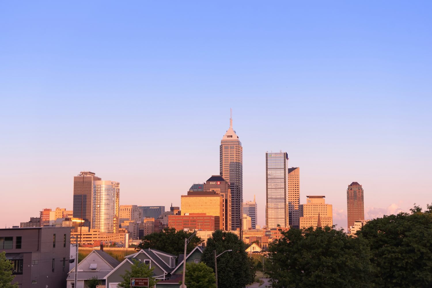 Indianapolis skyline at golden hour with the Salesforce Tower centered, BMO and Regions buildings nearby, and trees and homes in the foreground under a clear pastel sky.