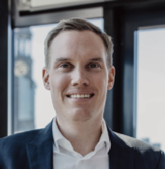 Porträt eines lächelnden Mannes in Business-Kleidung vor einem Fenster mit einer Uhr im Hintergrund.