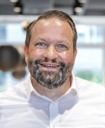 Porträt eines lächelnden Mannes mit braunen Haaren und Bart, der ein weißes Hemd trägt.