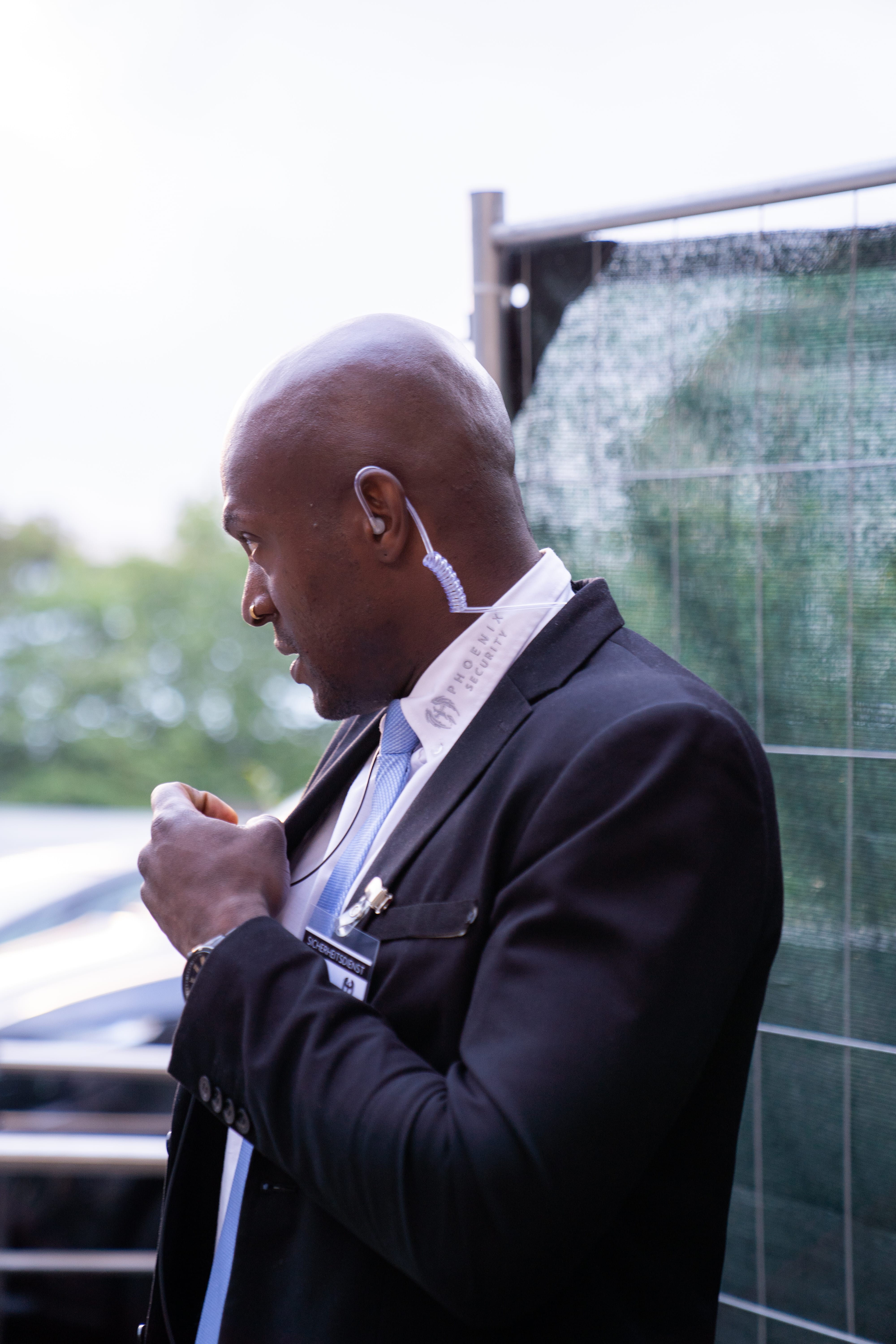 Seitliches Porträt eines Sicherheitsmanns mit Headset und schwarzem Anzug, der sich draußen nahe einem Zaun befindet.