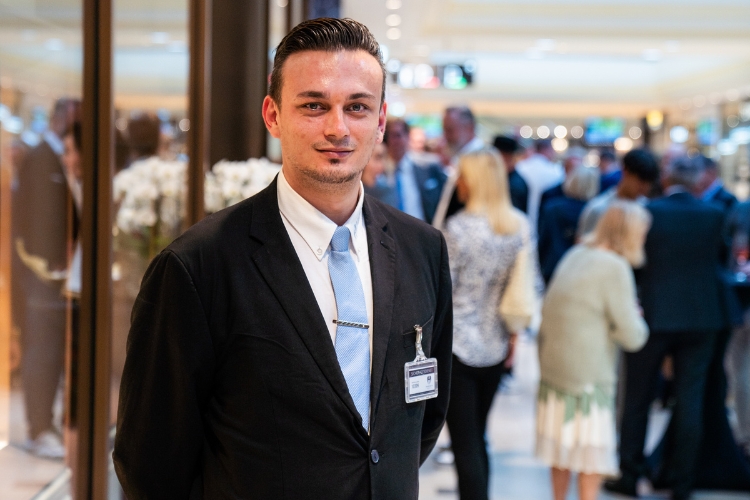 Mann in schwarzem Anzug mit hellblauer Krawatte und Namensschild steht in einem belebten VIP-Bereich.