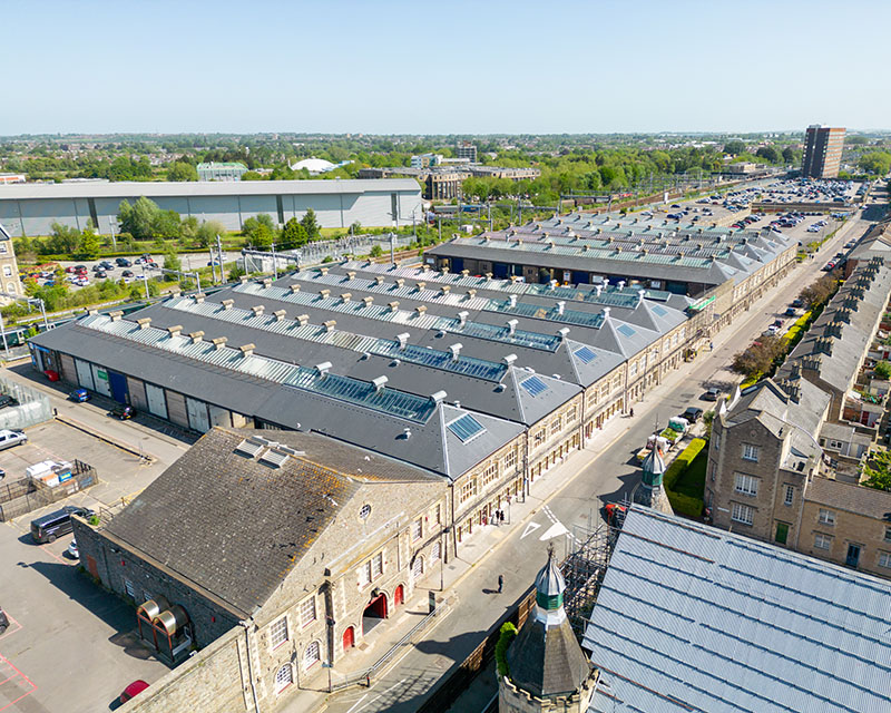 Aerial view of historic industrial-style buildings with pitched roofs featuring skylights, adjacent streets, and surrounding greenery under a clear sky.