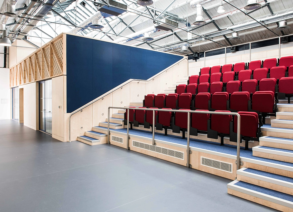 Modern lecture hall with tiered rows of red seats and light wood and blue panel walls under a glass ceiling.