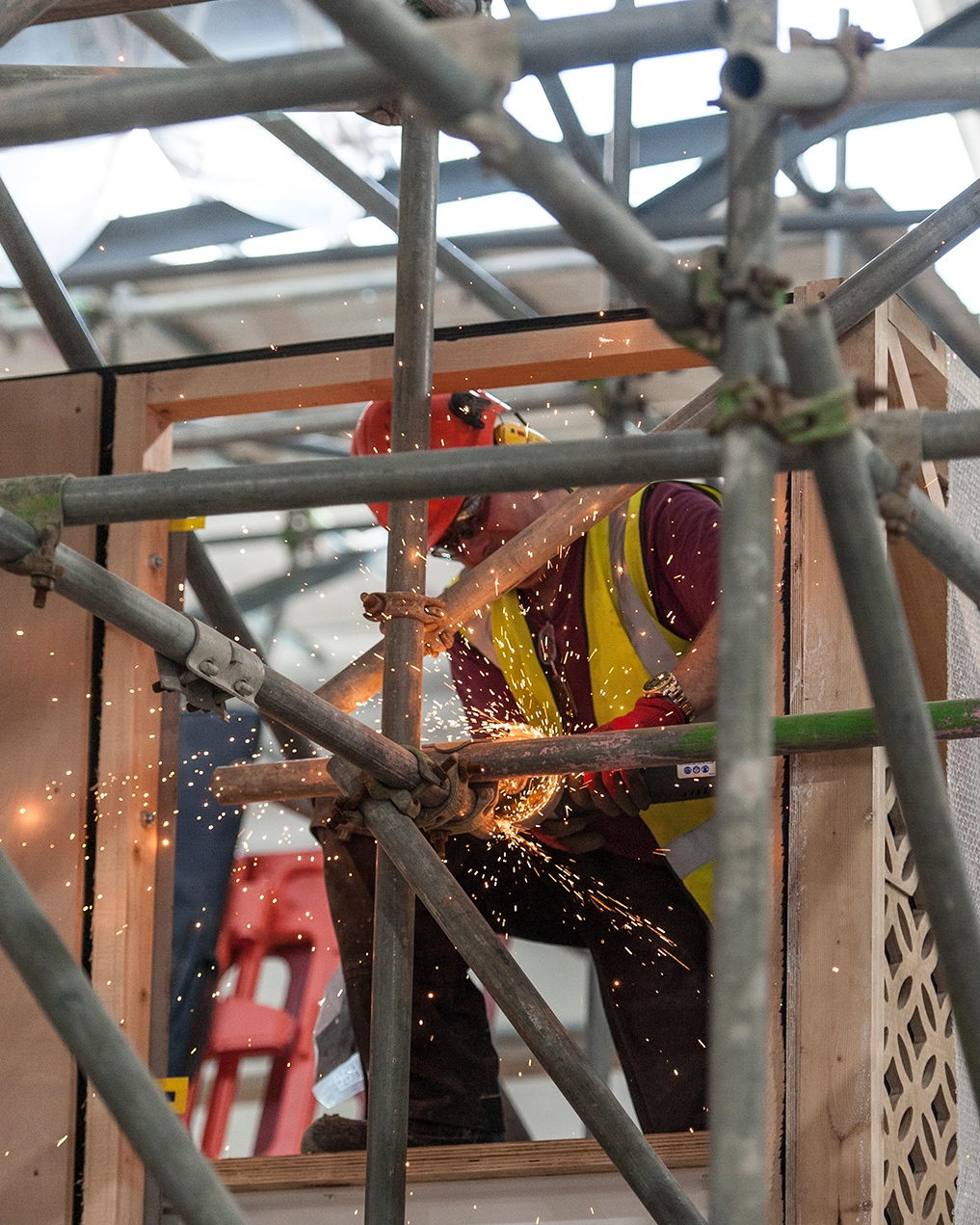 Construction worker wearing a red helmet and yellow safety vest cutting metal pipe with sparks flying, surrounded by scaffolding.