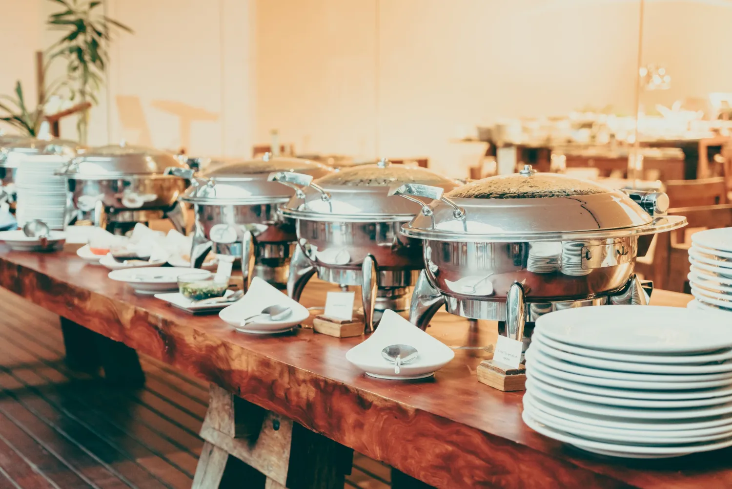 Buffet table with stainless steel chafing dishes, small plates, and serving spoons arranged neatly.