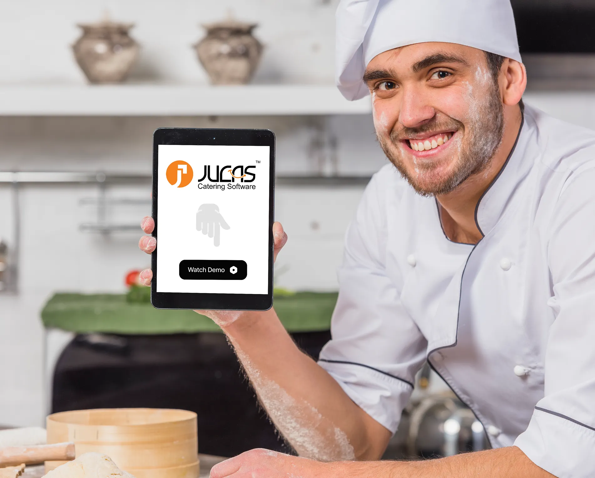 Smiling chef in a white uniform holding a tablet displaying the Jucas Catering Software logo and a Watch Demo button.