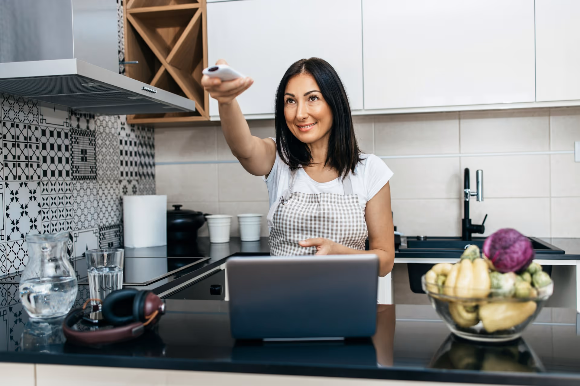 Woman in a modern kitchen wearing an apron, smiling as she points a remote control. 