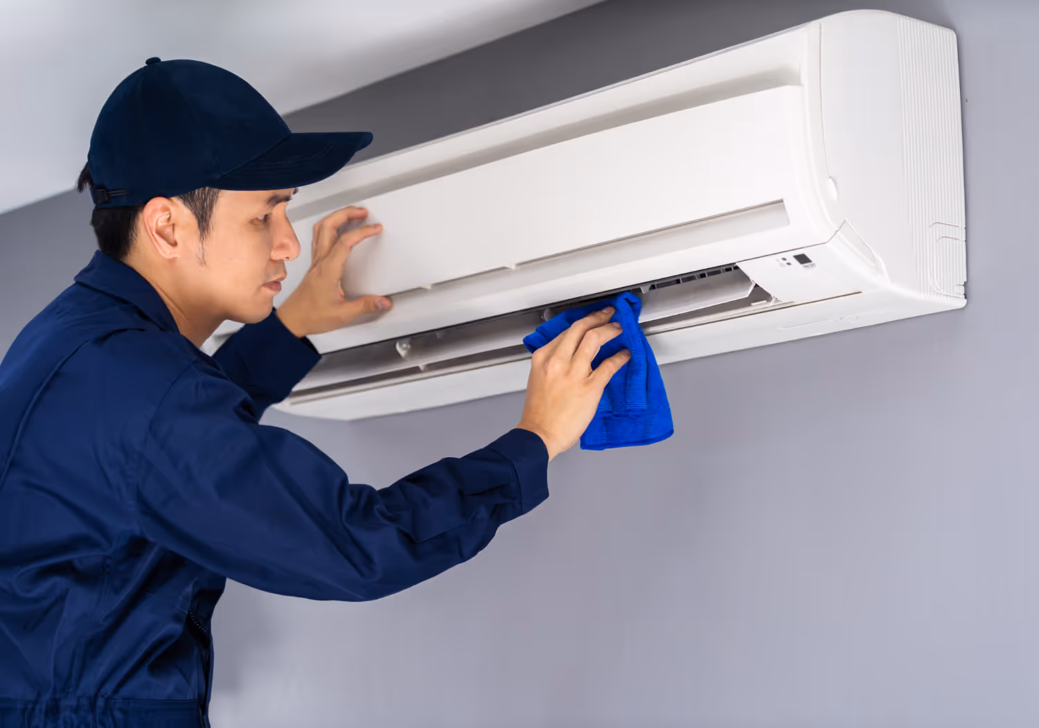 A technician in a blue uniform and cap cleans an air conditioning unit on a gray wall with a blue cloth