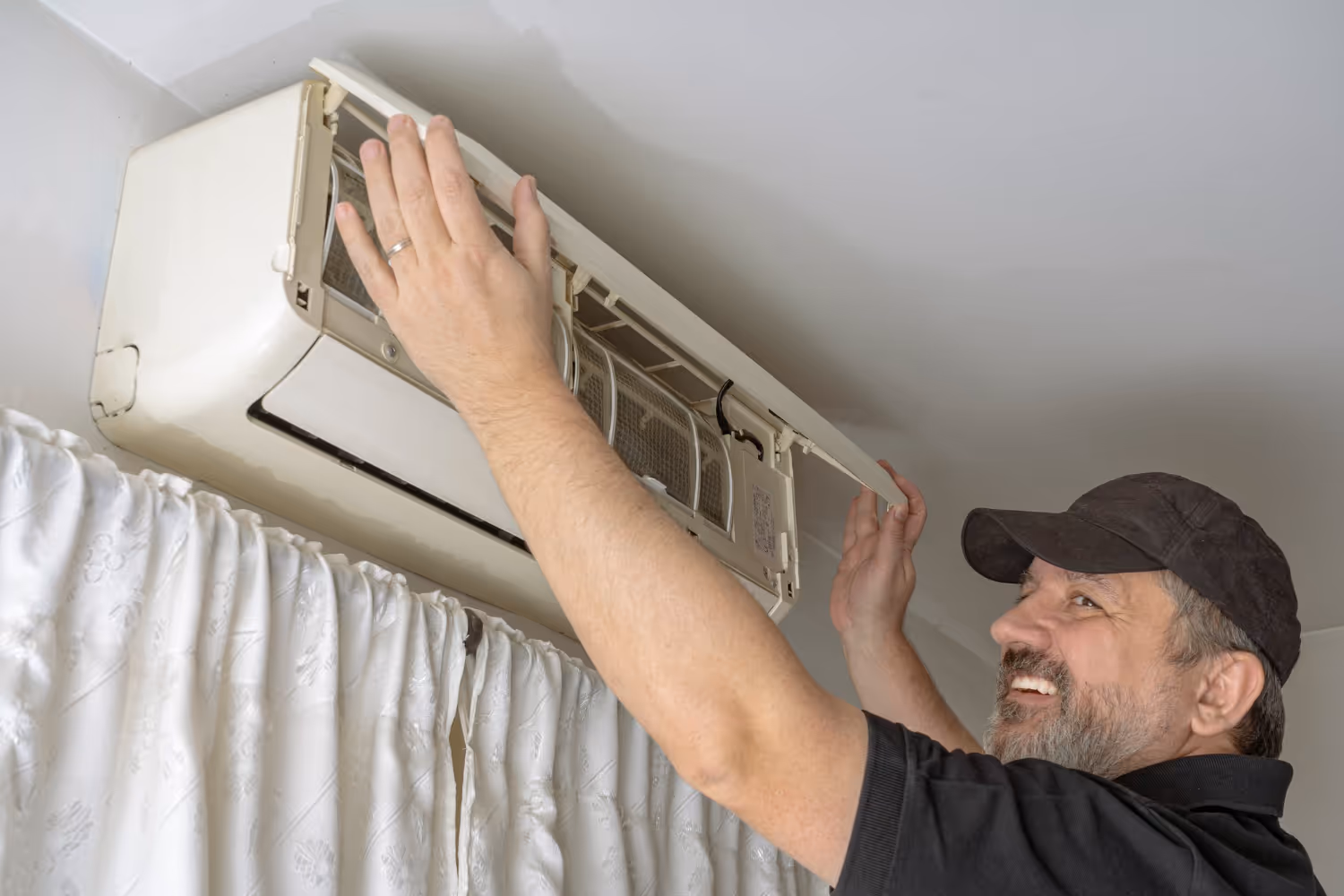 A man in a black cap smiles as he repairs a wall-mounted air conditioning unit, near white curtains.