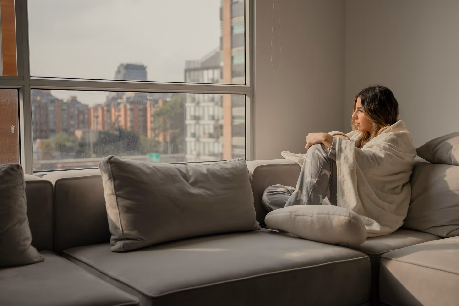 A woman wrapped in a blanket sits thoughtfully on a gray sofa by a window overlooking distant buildings. 